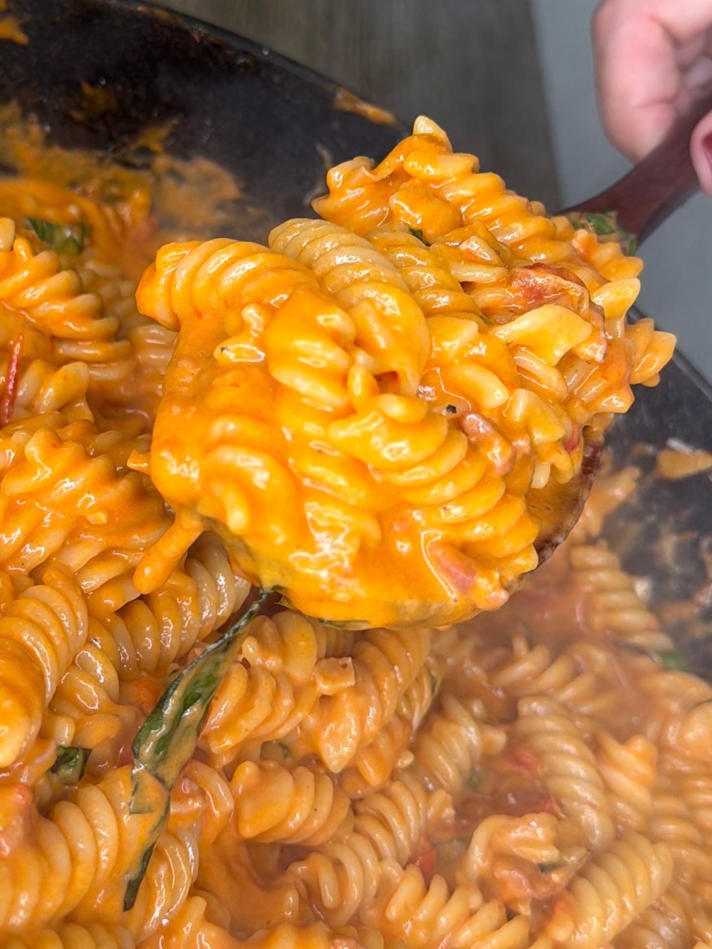 A close-up of a spoon lifting creamy tomato sauce-covered rotini pasta with basil from a pan.