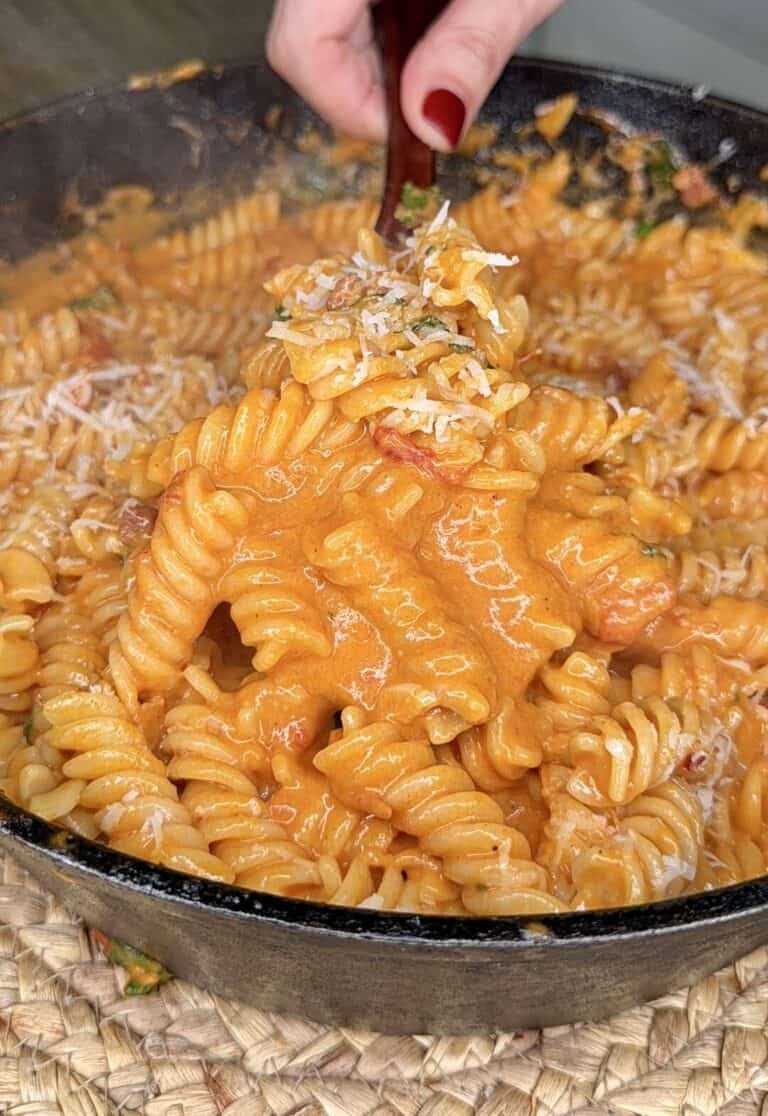 A hand holding a fork lifts creamy, tomato-sauced rotini pasta from a black skillet, topped with grated cheese.