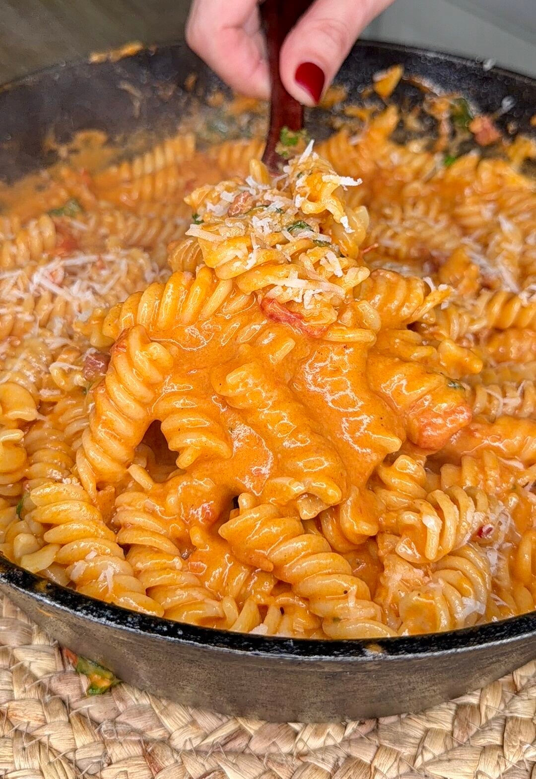 A hand holding a fork lifts creamy, tomato-sauced rotini pasta from a black skillet, topped with grated cheese.