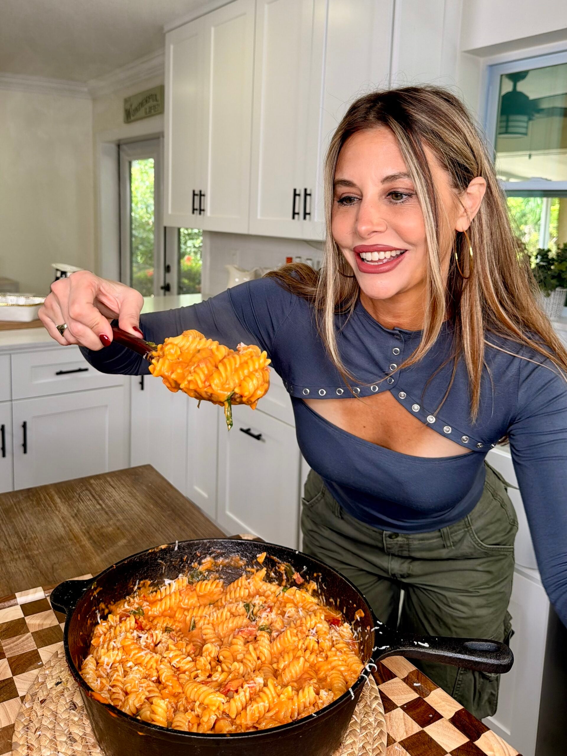 A smiling woman in a kitchen serves rotini pasta with tomato sauce from a skillet, holding a large spoon over a wooden table.