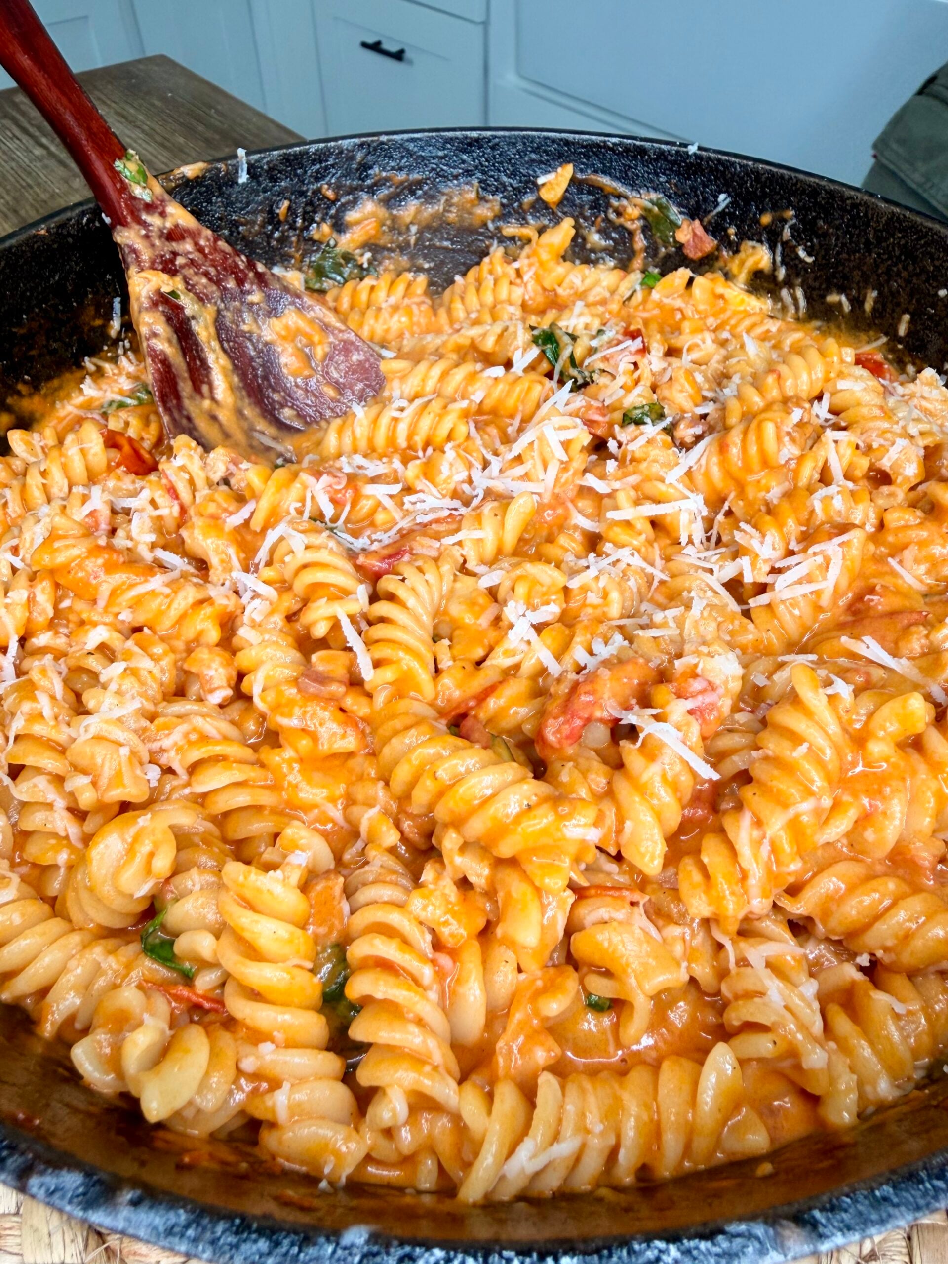 A skillet filled with creamy rotini pasta in a tomato sauce, topped with grated cheese and mixed with a wooden spoon.