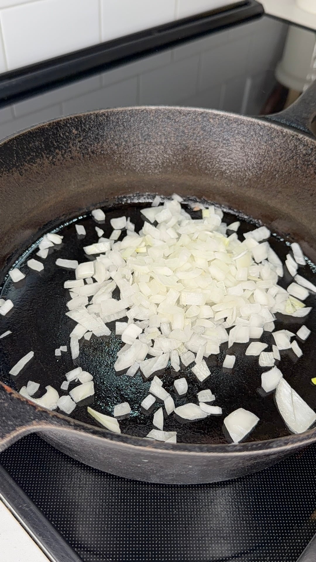 Chopped onions cooking in a black cast iron skillet on a stovetop, with a white tiled backsplash visible in the background.