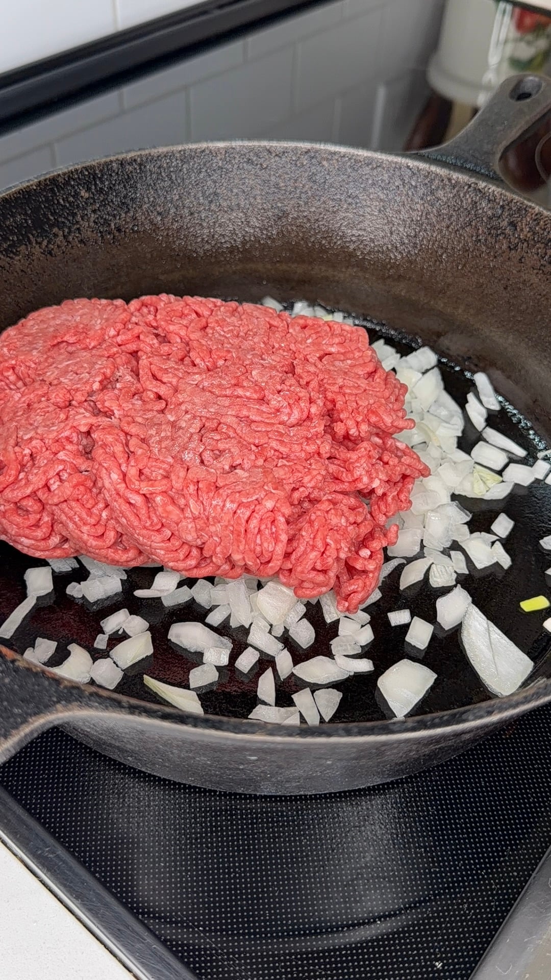 Raw ground beef and chopped onions in a black cast iron skillet on a stovetop, ready to be cooked.