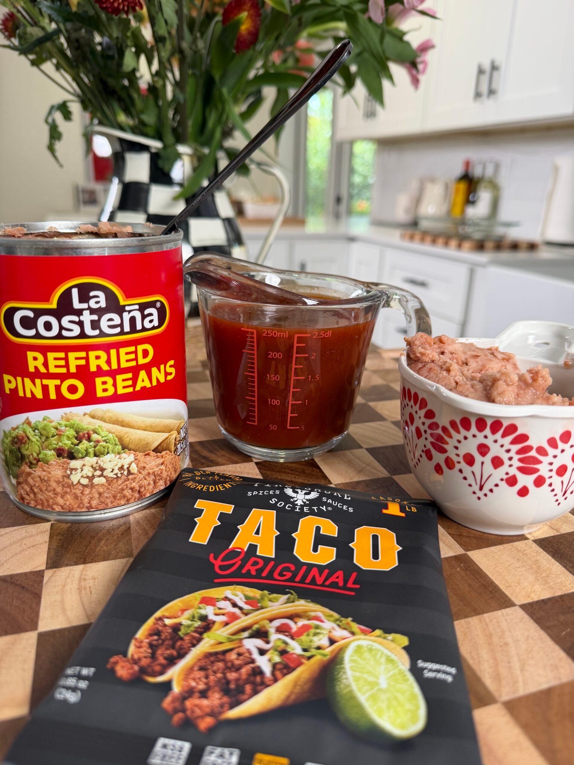 A can of refried pinto beans, a measuring cup with sauce, a taco seasoning packet, and a small bowl with beans on a wooden counter in a bright kitchen with flowers in the background.