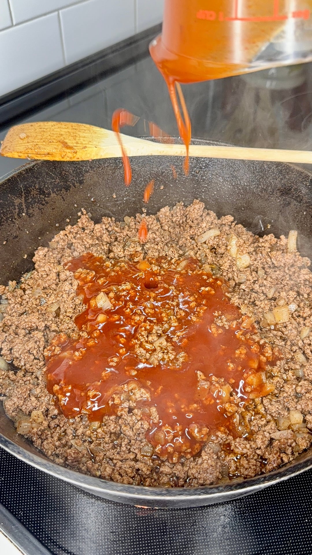 Ground beef and onions are cooking in a skillet while red sauce is being poured in from a measuring cup. A wooden spatula rests on the stove nearby. Steam rises from the pan.