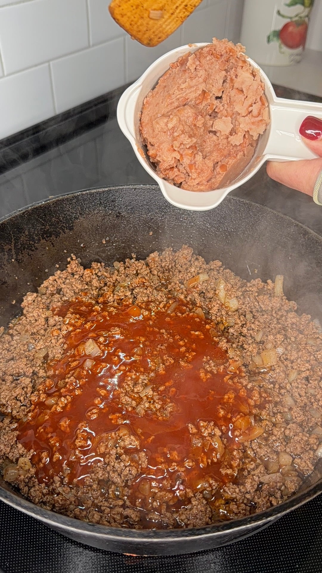 Ground beef cooking in a skillet with sauce, while a person holds a bowl of refried beans above the pan, preparing to add them.