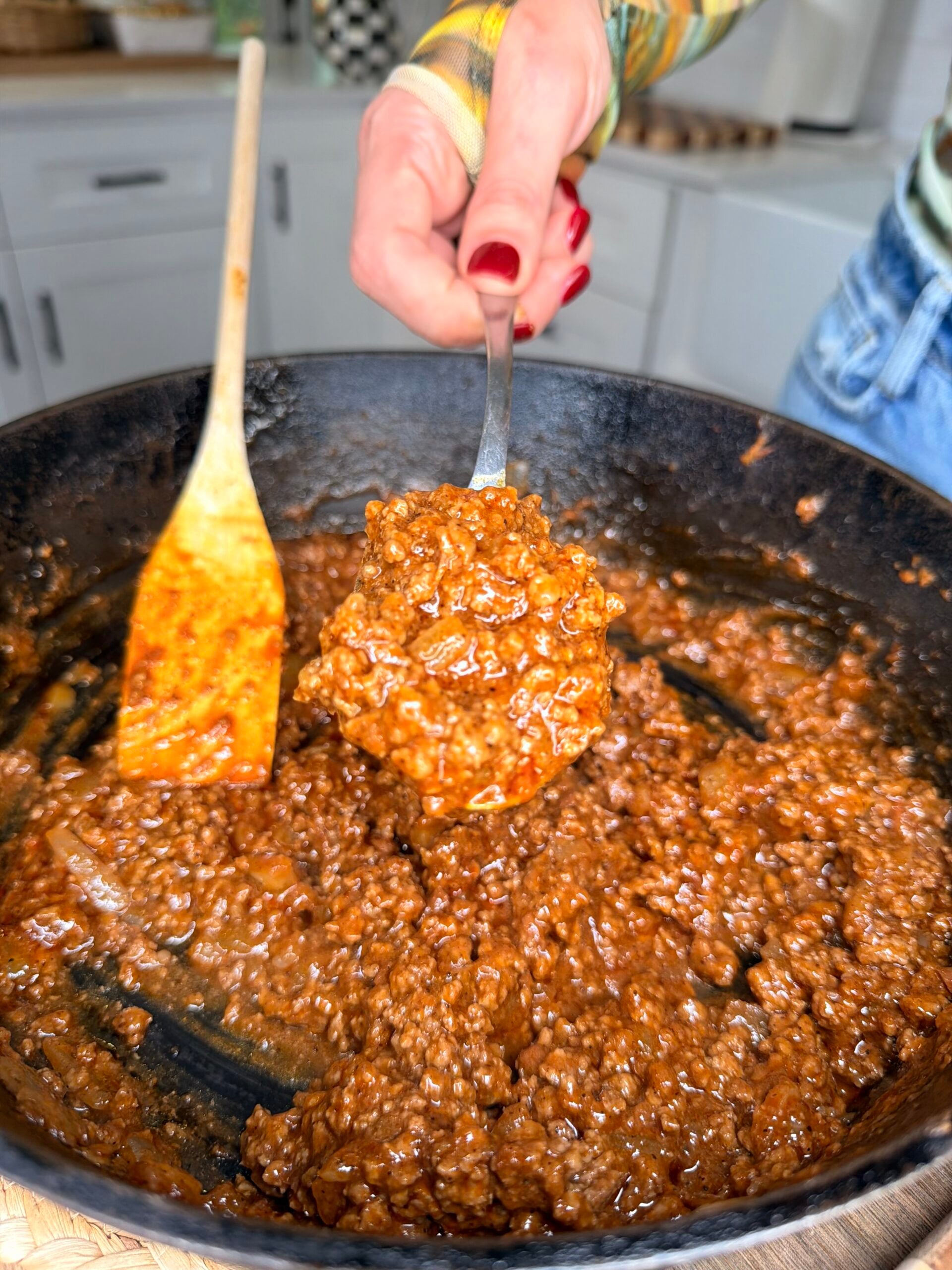 A person holds a spoonful of cooked, saucy ground beef over a skillet, with a wooden spatula resting inside the pan in a kitchen setting.