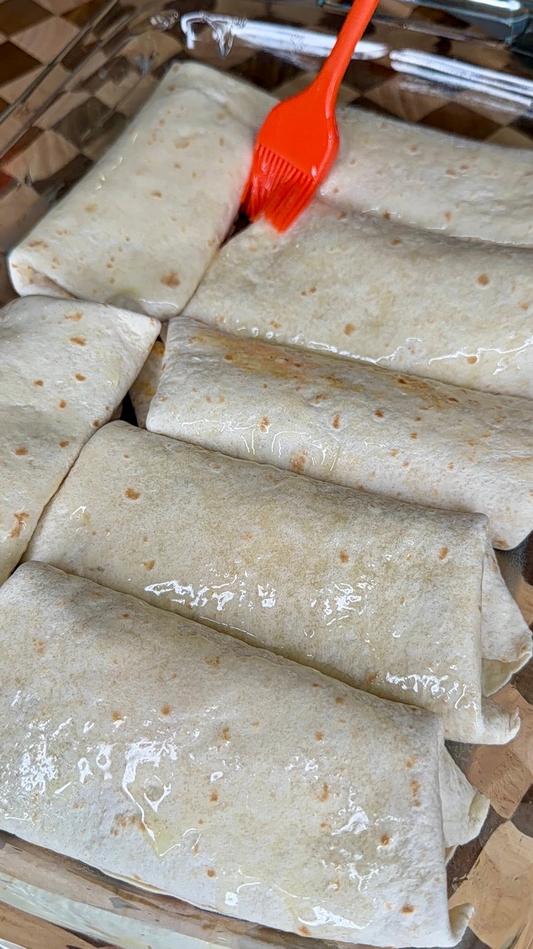 A close-up of several rolled tortillas in a glass baking dish, with an orange brush being used to spread oil or butter on top.