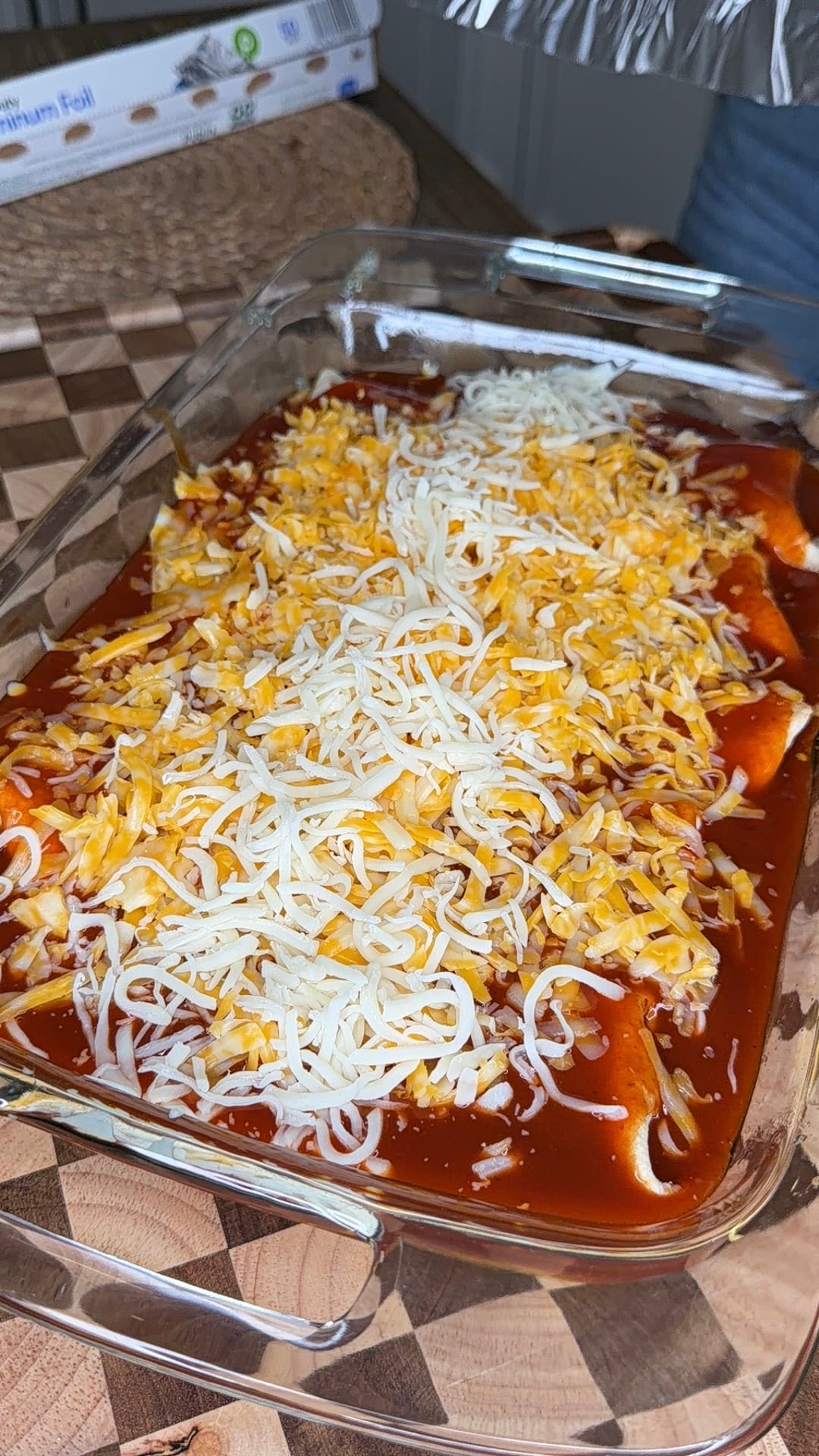 A glass baking dish filled with enchiladas topped with red sauce and a generous layer of shredded cheese, sitting on a wooden surface.