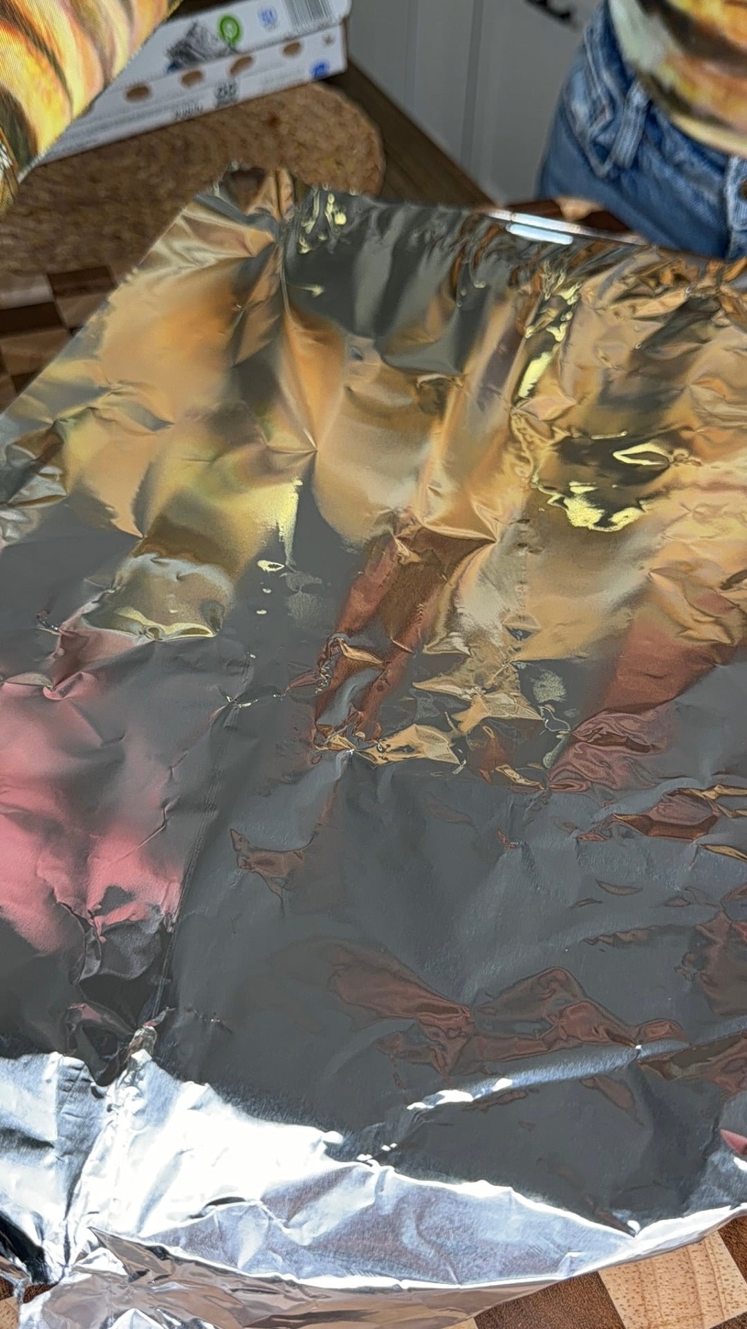 A tray covered with crinkled aluminum foil sits on a countertop; part of a person in jeans and a colorful shirt is visible beside it.