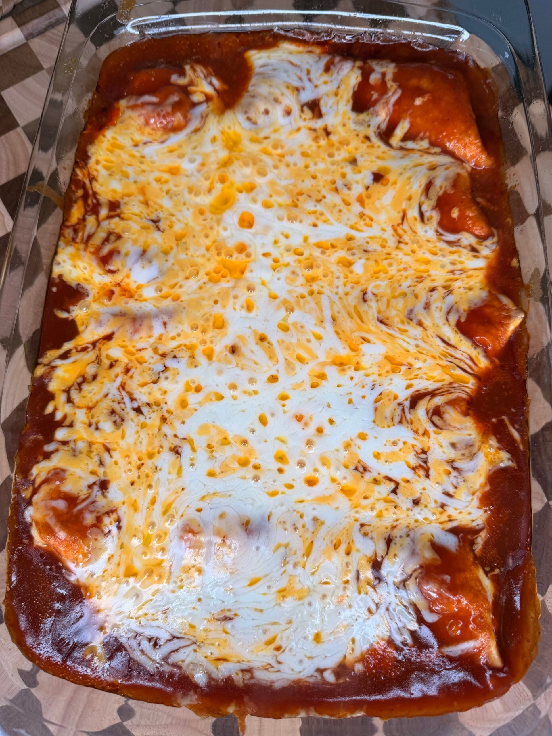 A glass baking dish filled with enchiladas topped with melted cheese and red enchilada sauce on a wooden surface.