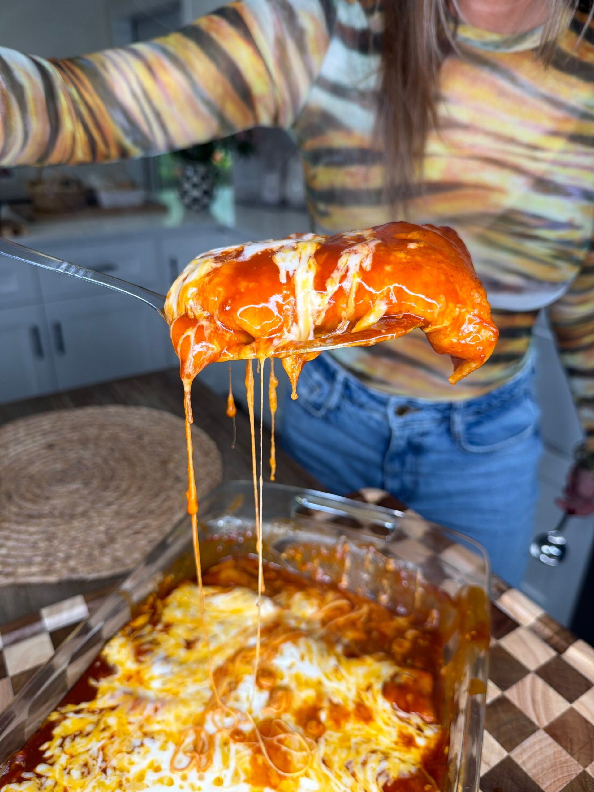 A person in a striped shirt holds a cheesy, saucy enchilada with a fork above a glass baking dish filled with enchiladas on a kitchen counter.