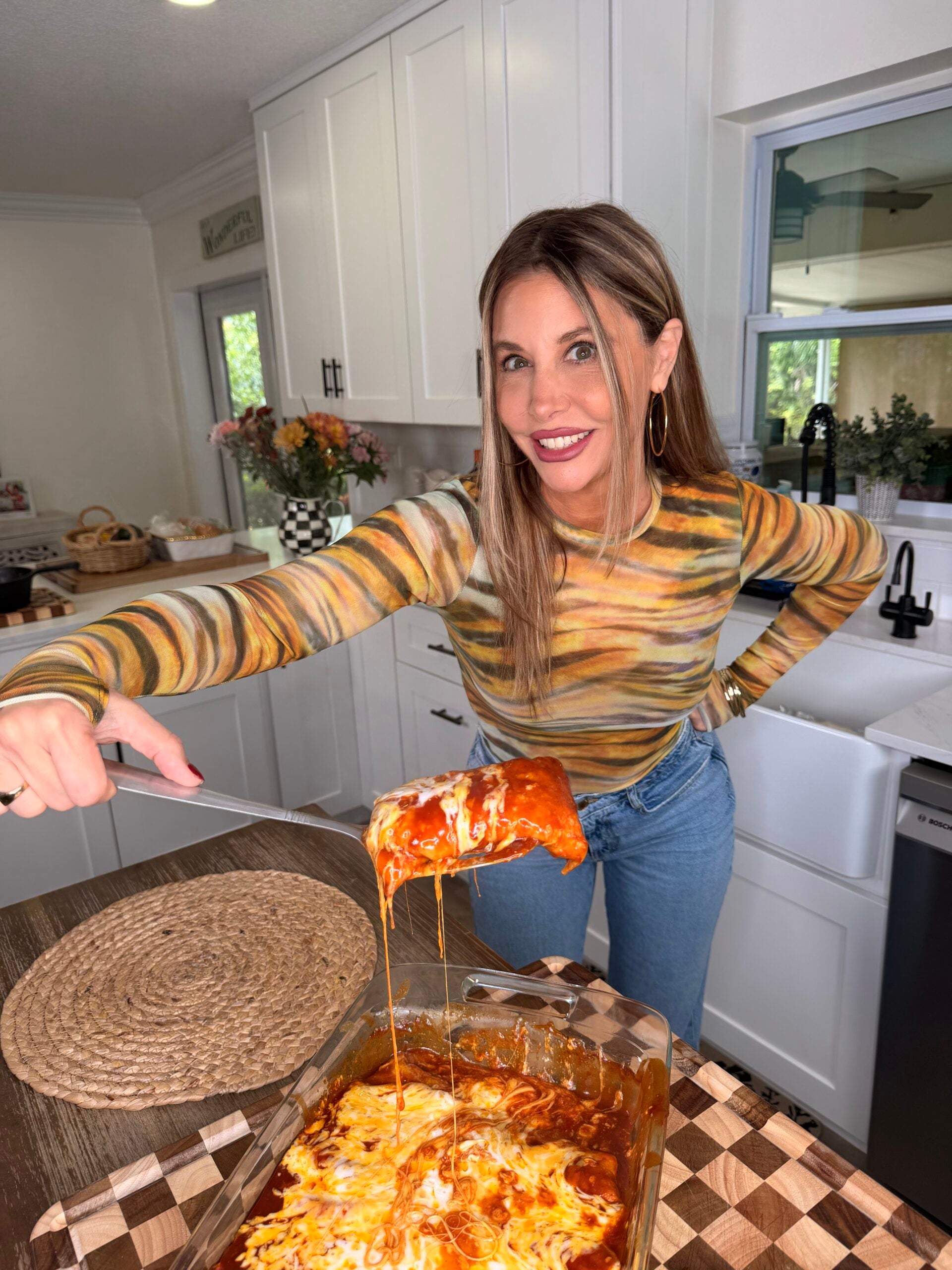 A woman smiles while serving a cheesy baked pasta dish from a glass pan in a bright kitchen, wearing a striped long-sleeve shirt and jeans.