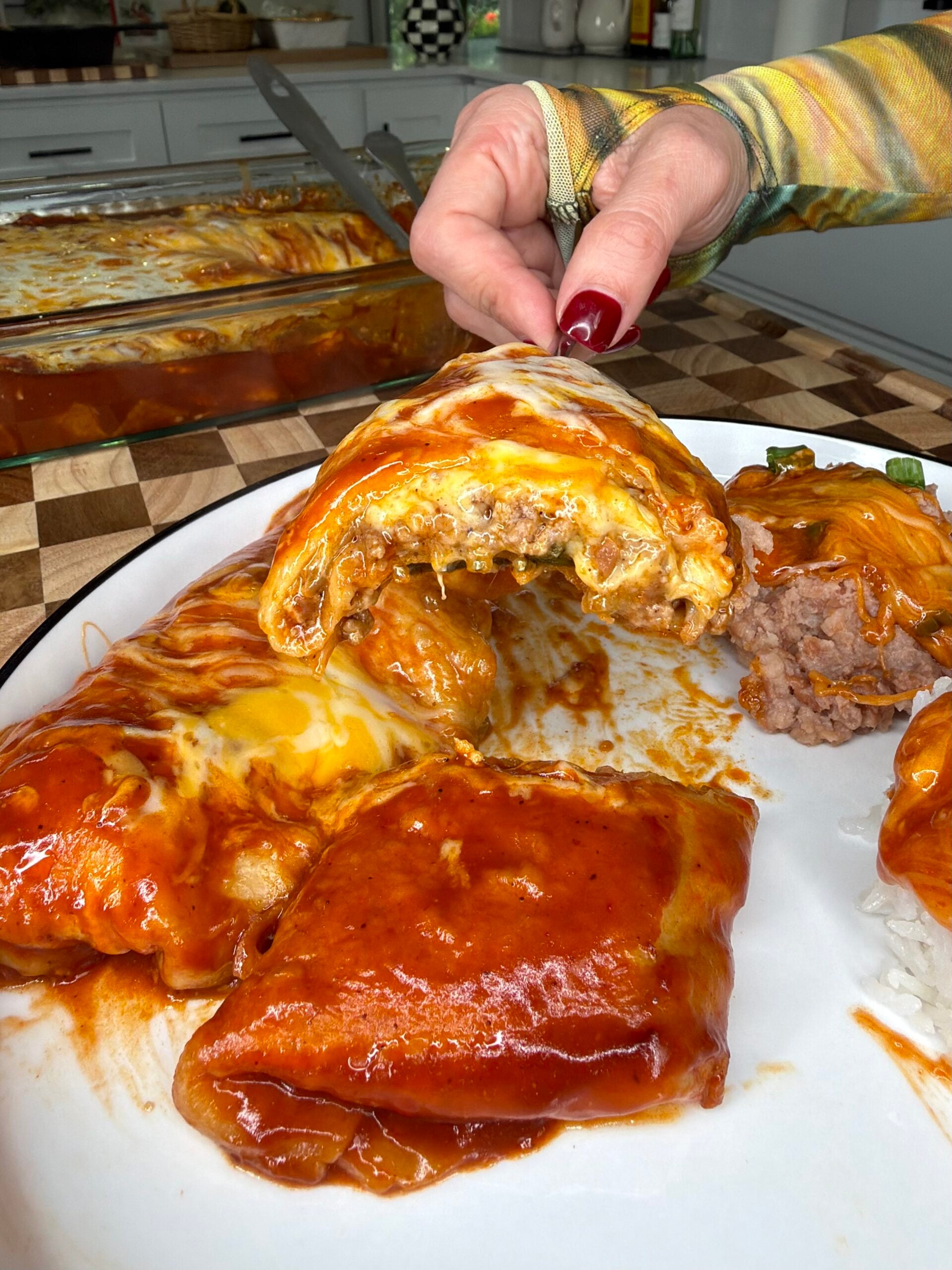A hand holds a cheesy, saucy enchilada over a plate with more enchiladas and refried beans. The background shows a baking dish with extra enchiladas on a checkered kitchen counter.