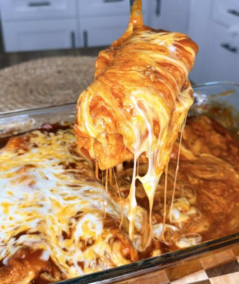 A cheesy, saucy enchilada is being lifted from a baking dish, with melted cheese stretching, in a bright kitchen setting.