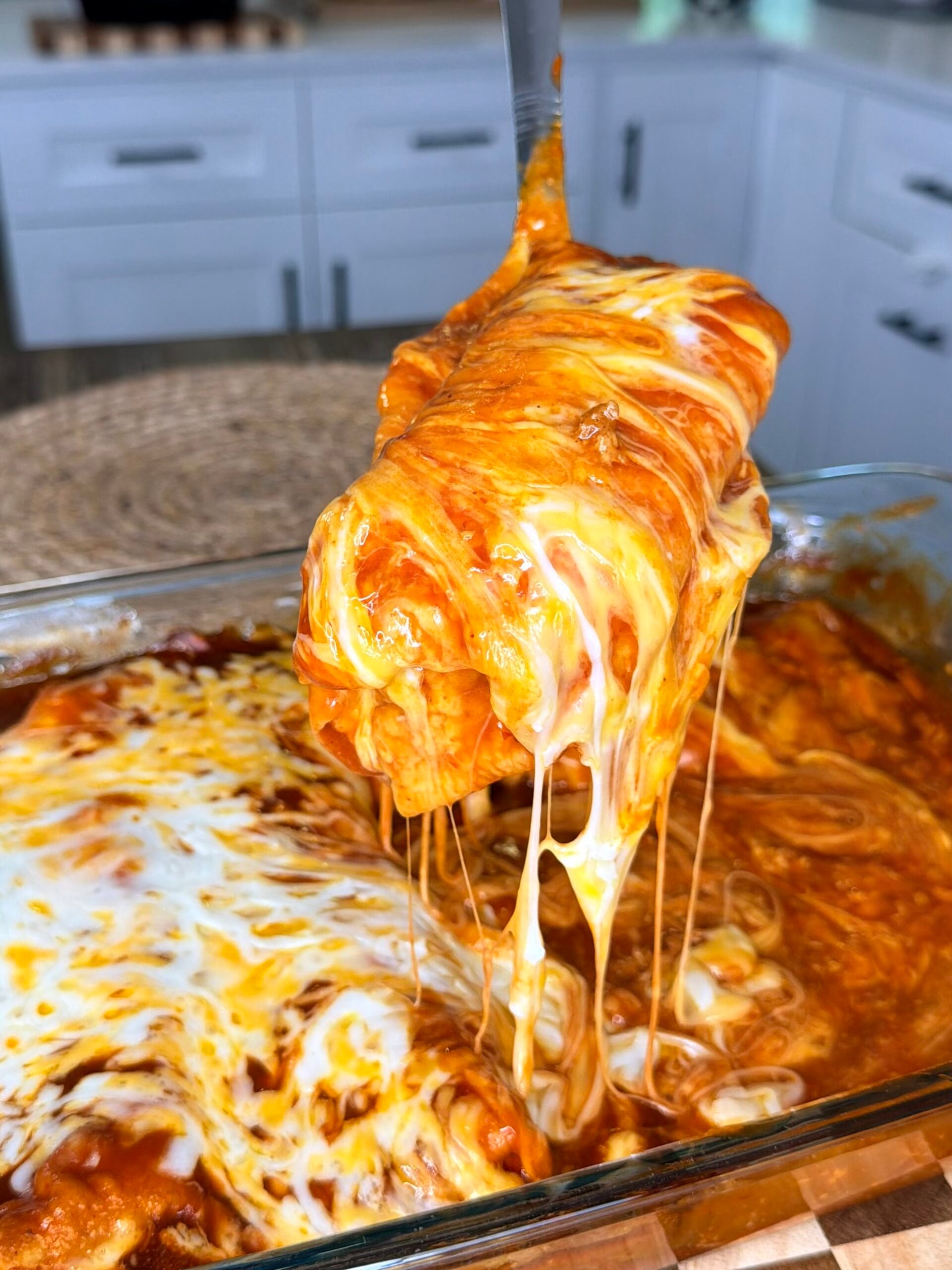 A cheesy enchilada is being lifted from a glass baking dish, with melted cheese stretching, in a kitchen setting.