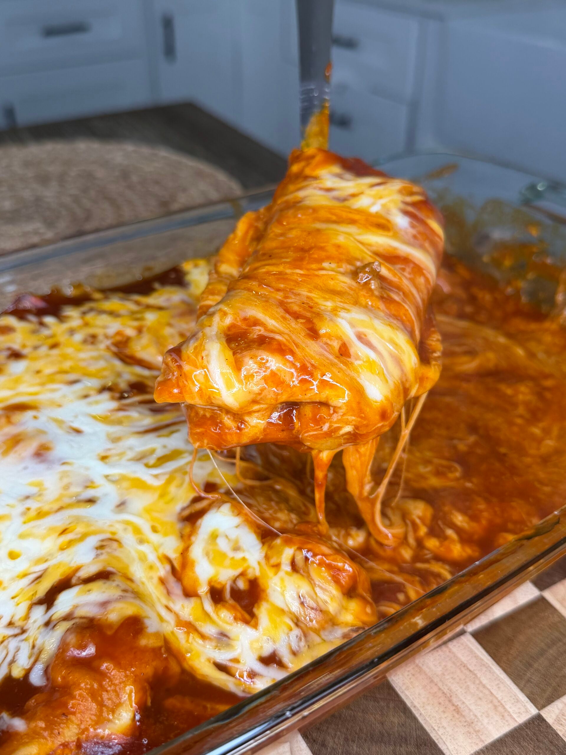 A close-up of a cheesy, saucy enchilada being lifted from a glass baking dish filled with melted cheese and red sauce.