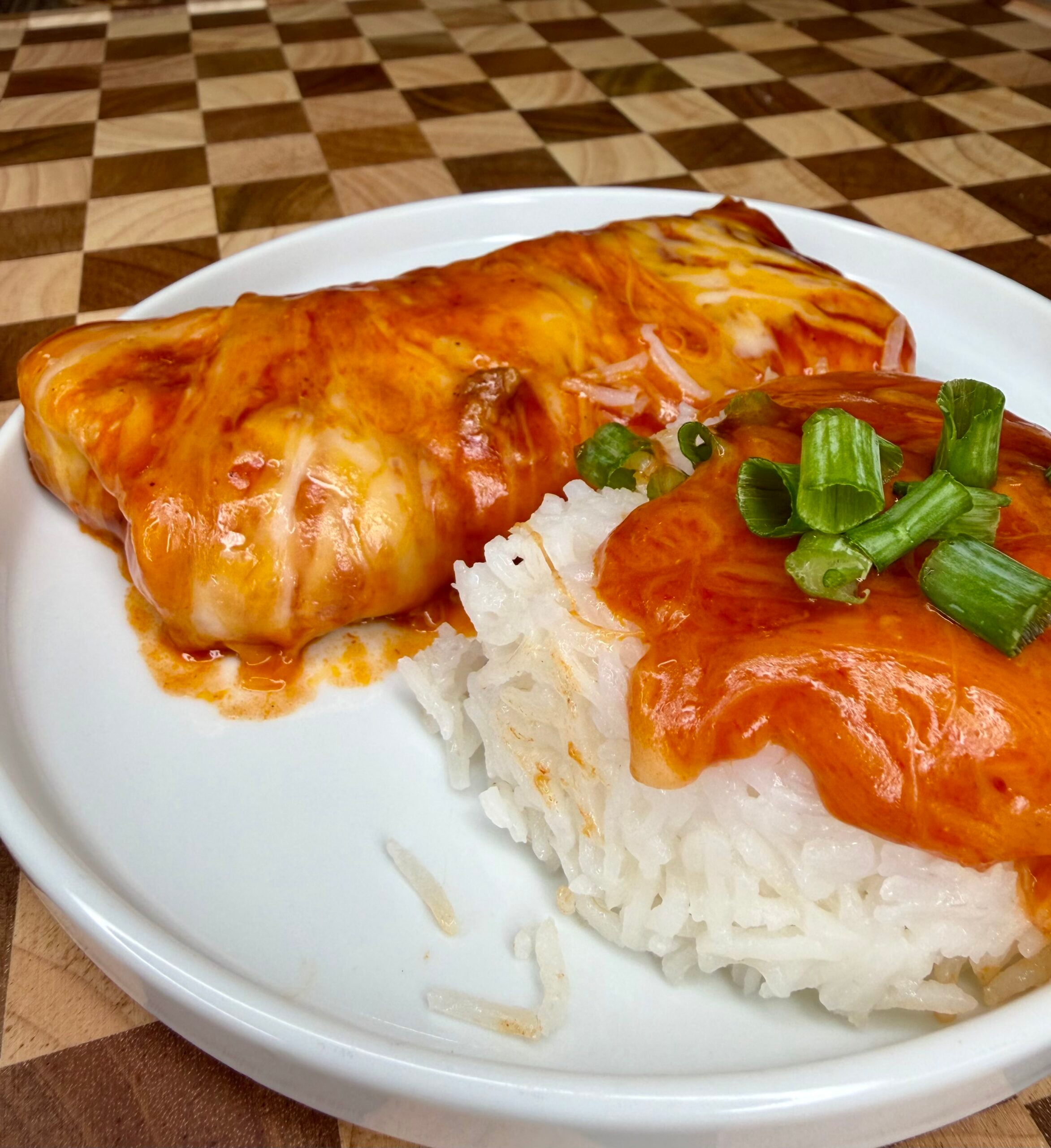 A plate with an enchilada covered in red sauce and melted cheese next to a mound of white rice topped with sauce and chopped green onions, on a wooden checkered surface.