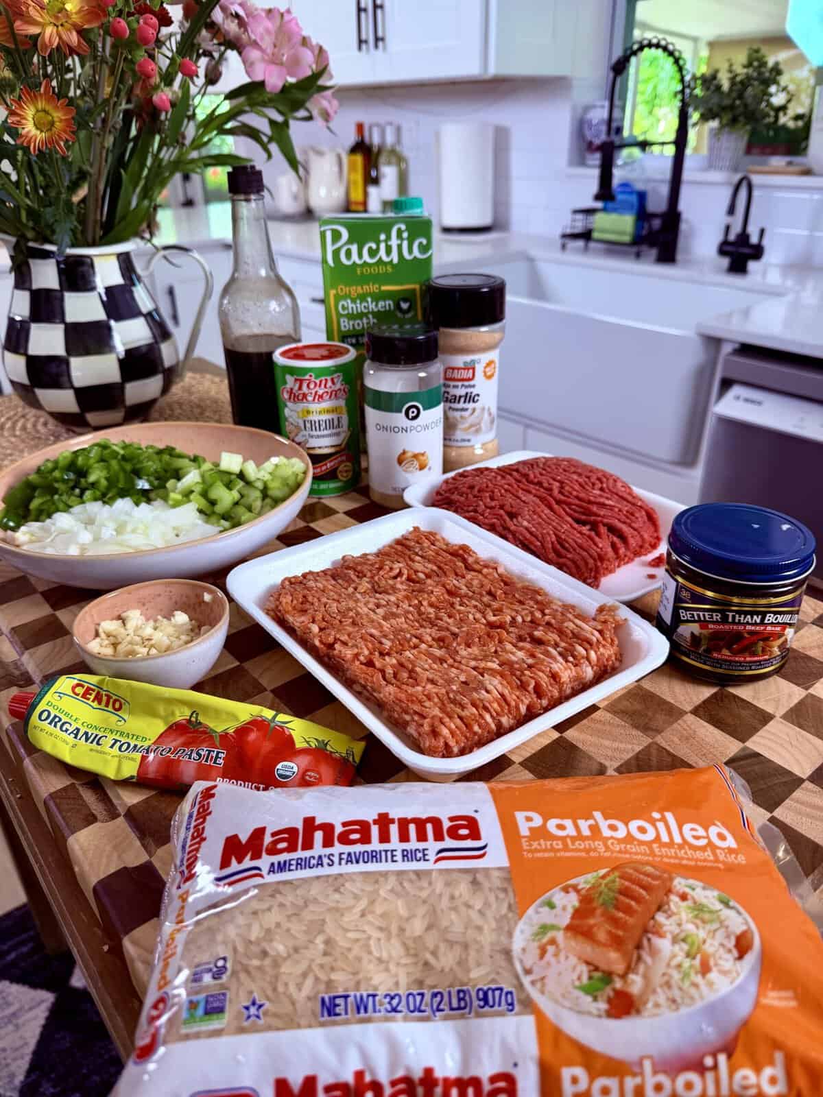 A kitchen counter with ingredients for a meal, including ground meats, rice, celery, green onions, garlic, seasonings, broth, tomato paste, and flowers in the background.