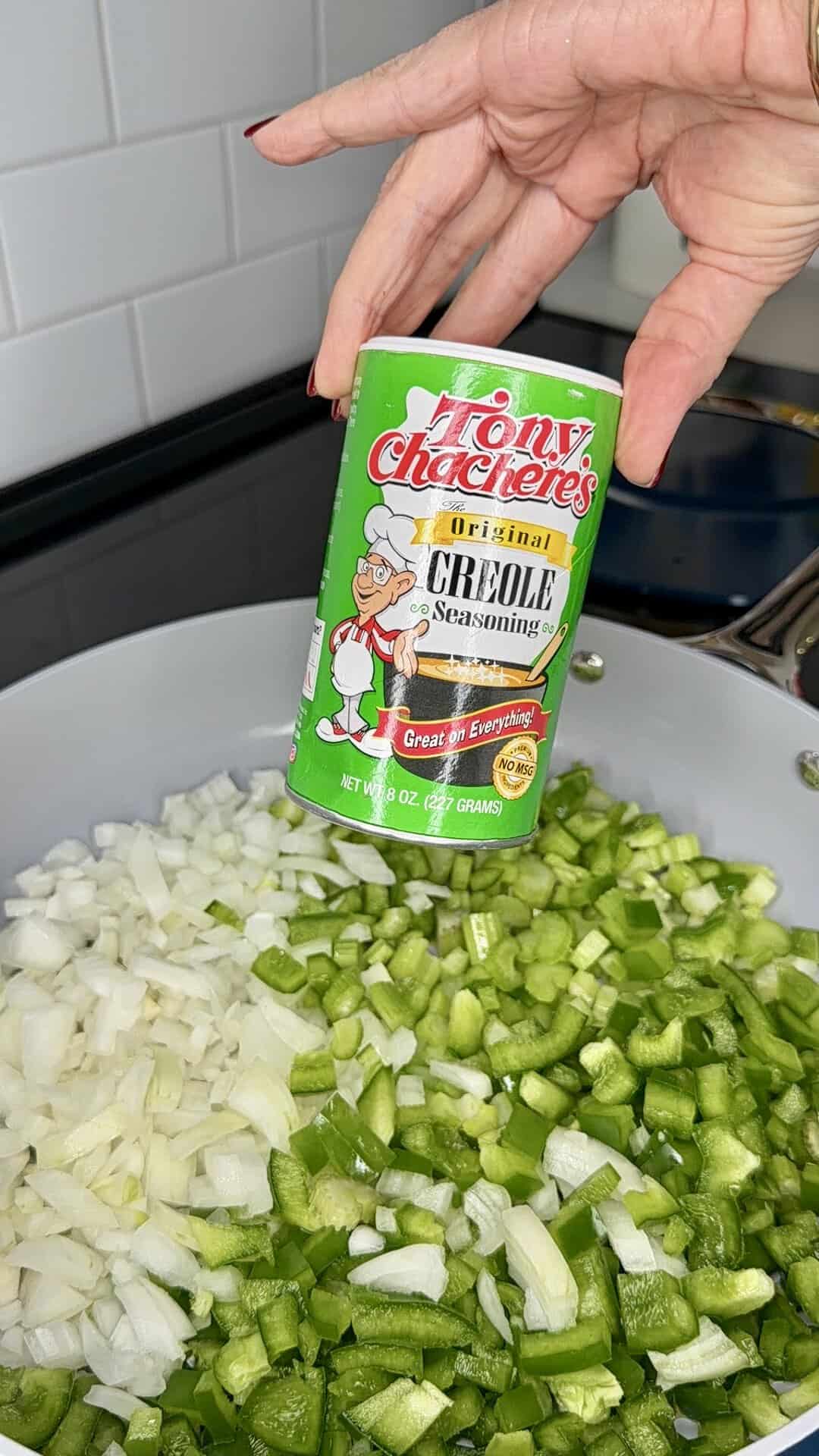 A hand holds a can of Tony Chacheres Creole Seasoning above chopped onions and green bell peppers in a pan on a stovetop.