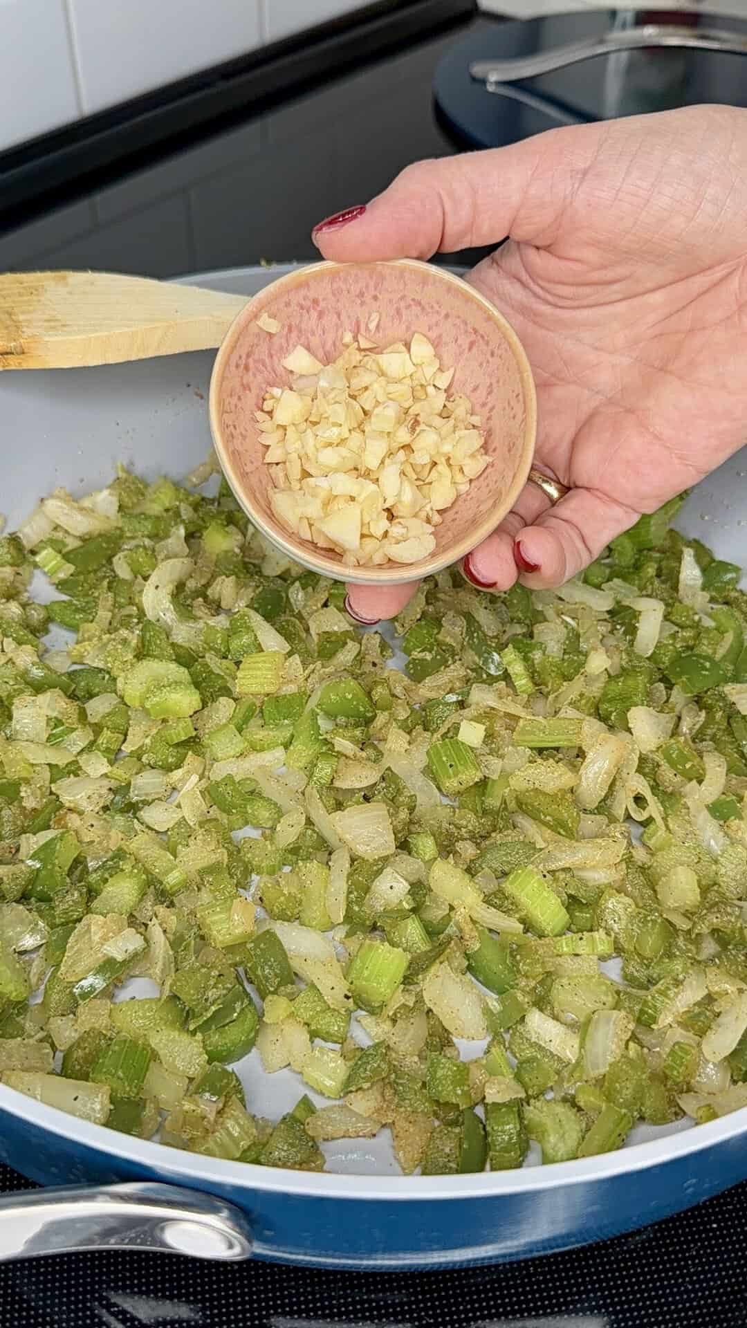 A hand holds a small bowl of chopped garlic over a pan filled with sautéed celery and onions on a stovetop, ready to add the garlic.