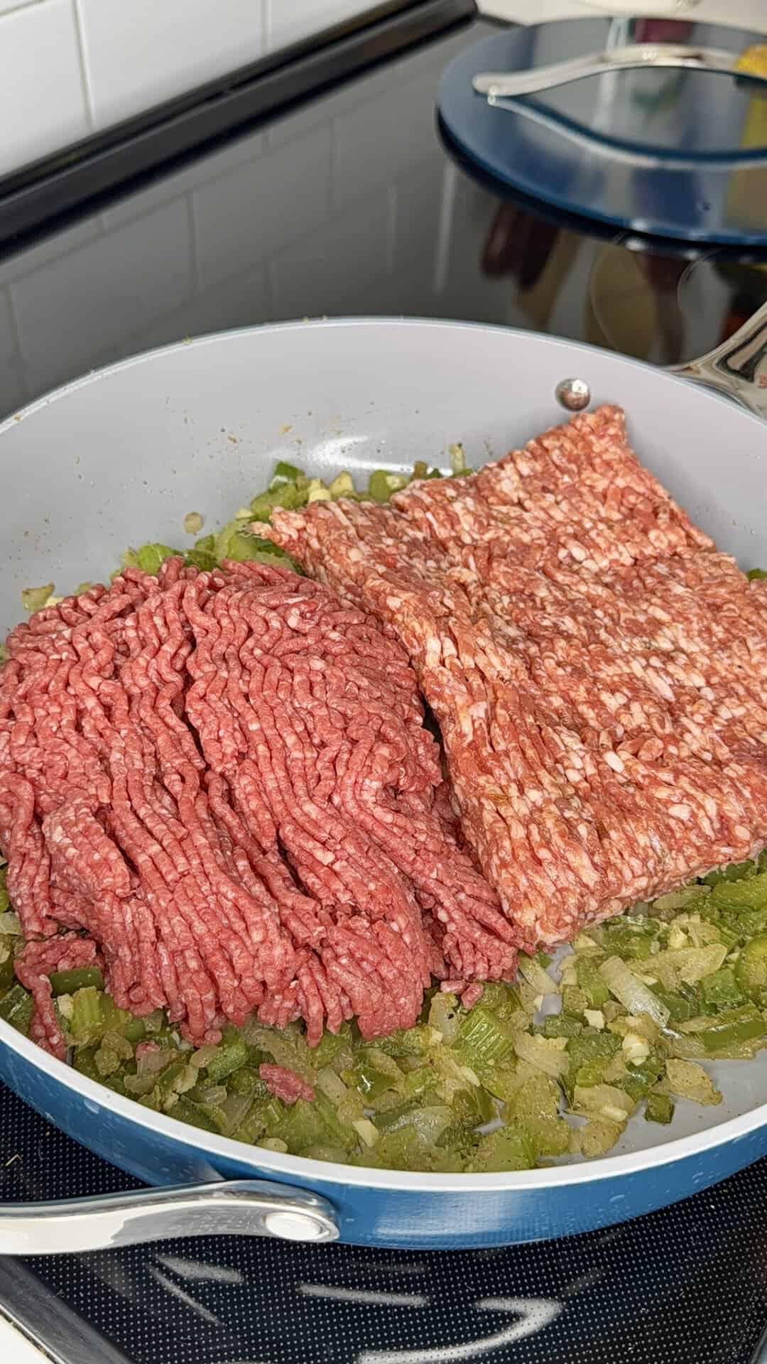 A frying pan on a stove contains chopped green peppers and onions, topped with raw ground beef and ground sausage, ready to cook.