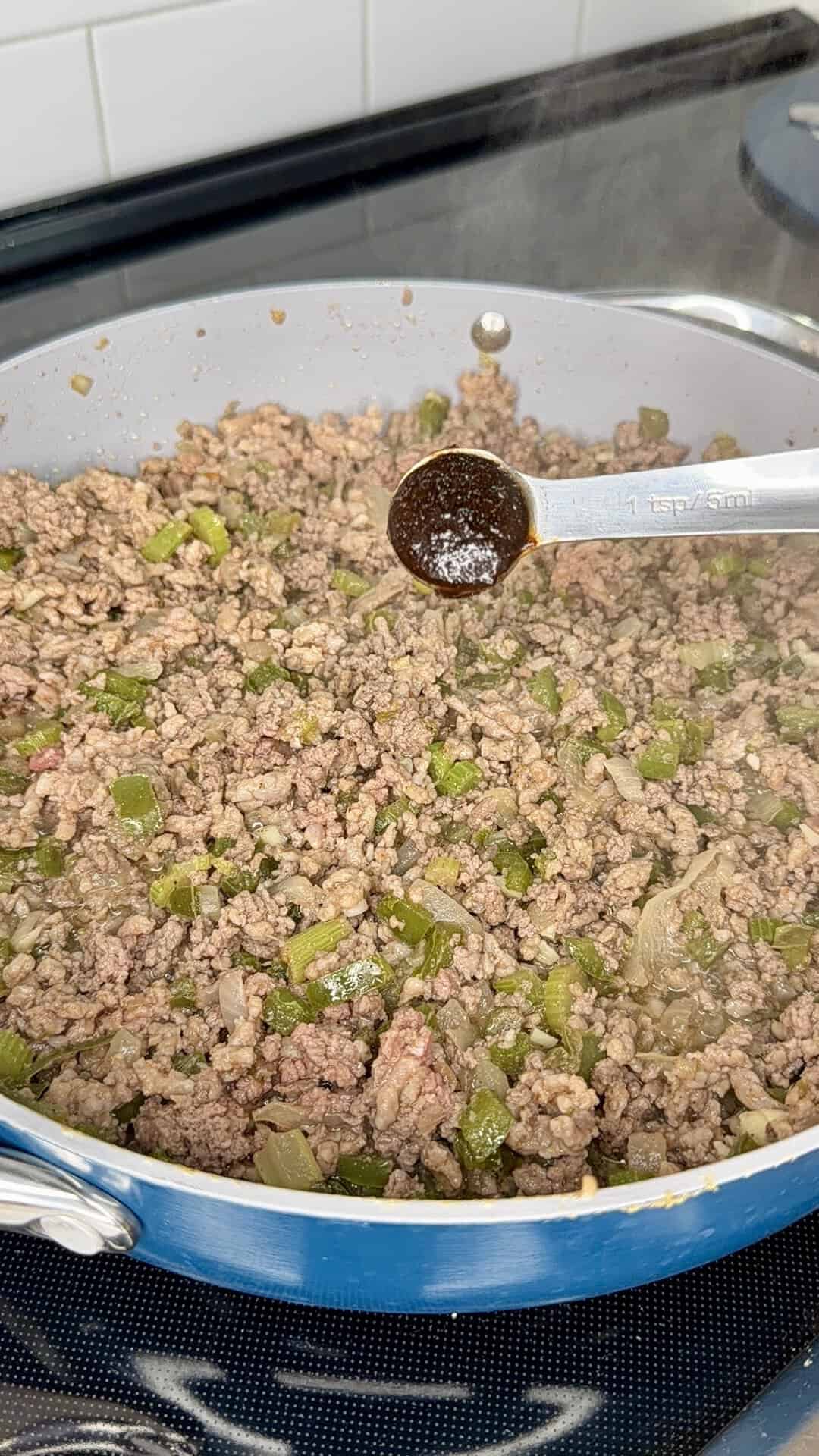 Ground beef, onions, and green peppers are cooking in a skillet on a stovetop. A metal measuring spoon filled with seasoning is held above the mixture, ready to be added. Steam is visible from the hot pan.