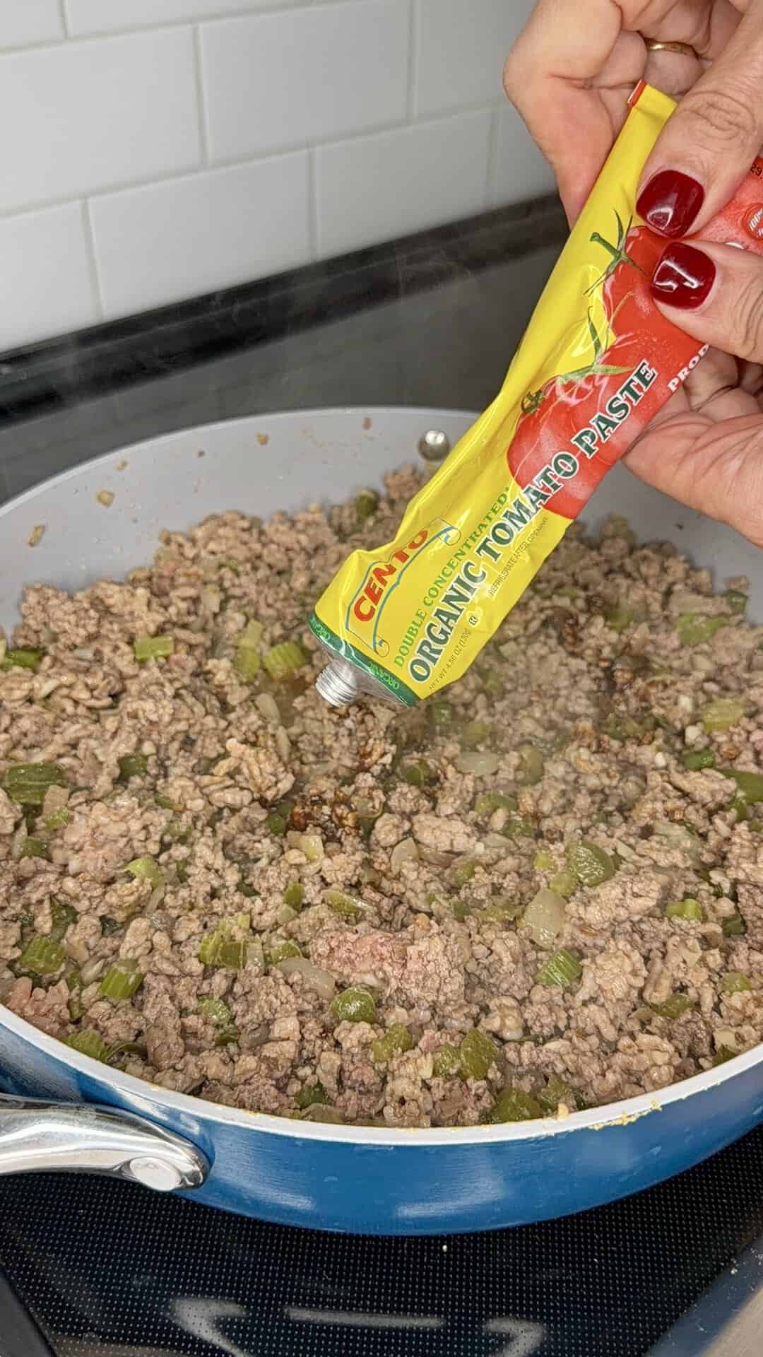 A hand squeezes organic tomato paste from a yellow tube into a pan of cooked ground beef and chopped green vegetables on a stovetop.