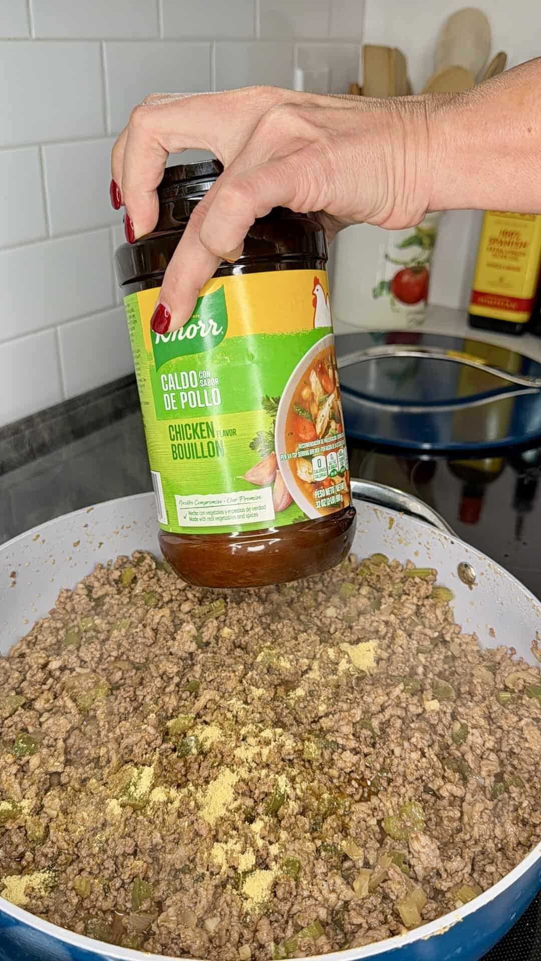 A hand sprinkles Knorr chicken bouillon seasoning from a jar over cooked ground beef in a skillet on a stovetop, with steam rising from the meat.