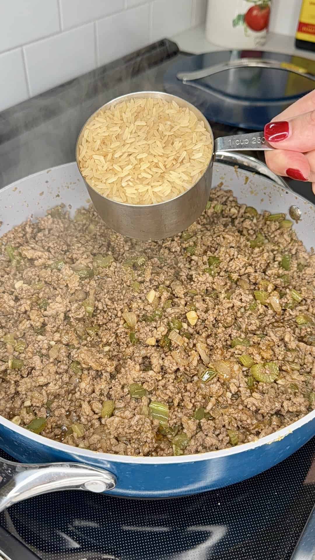 A hand holds a measuring cup of uncooked rice above a skillet filled with cooked ground beef and diced vegetables on a stovetop.