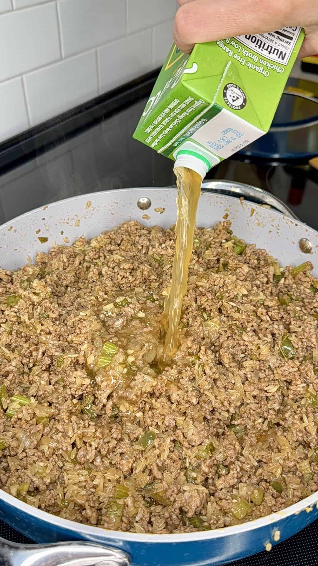 A hand pours broth from a green carton into a skillet filled with browned ground meat, rice, and chopped vegetables on a stovetop.
