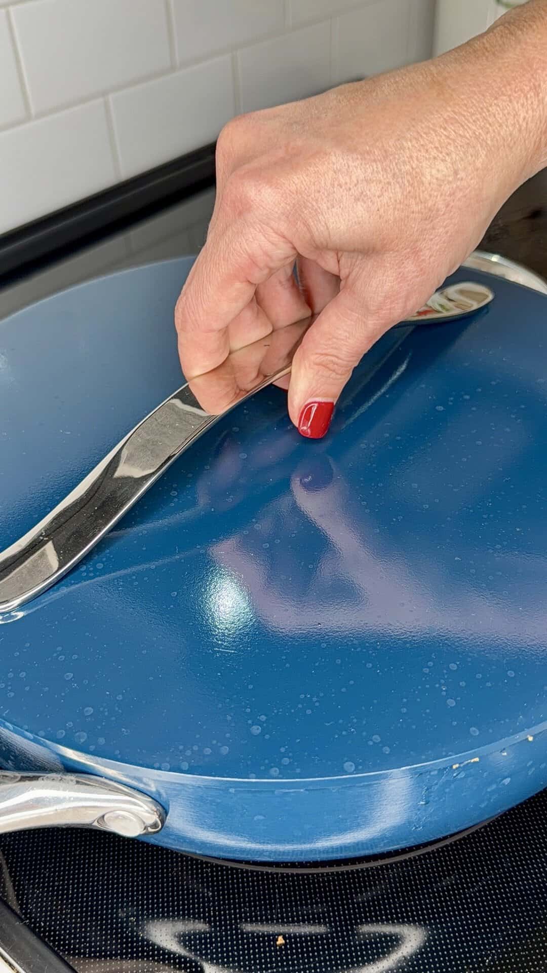A hand with red nail polish lifts the silver handle of a blue pot lid on a stovetop, with white tiled backsplash visible in the background.