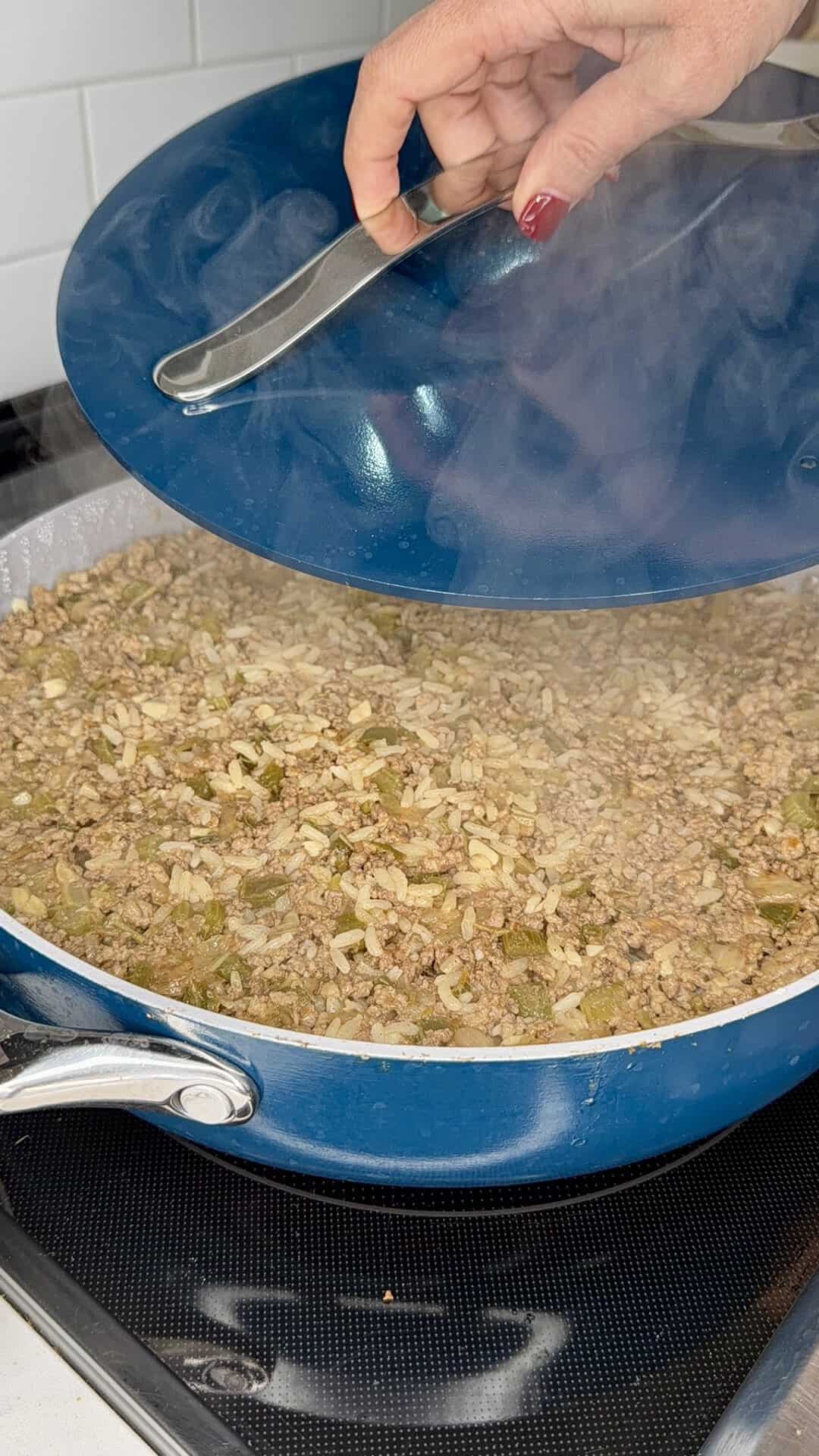 A hand lifts the lid off a blue skillet filled with cooked ground beef, onions, and green peppers on a stovetop, with steam rising from the pan.