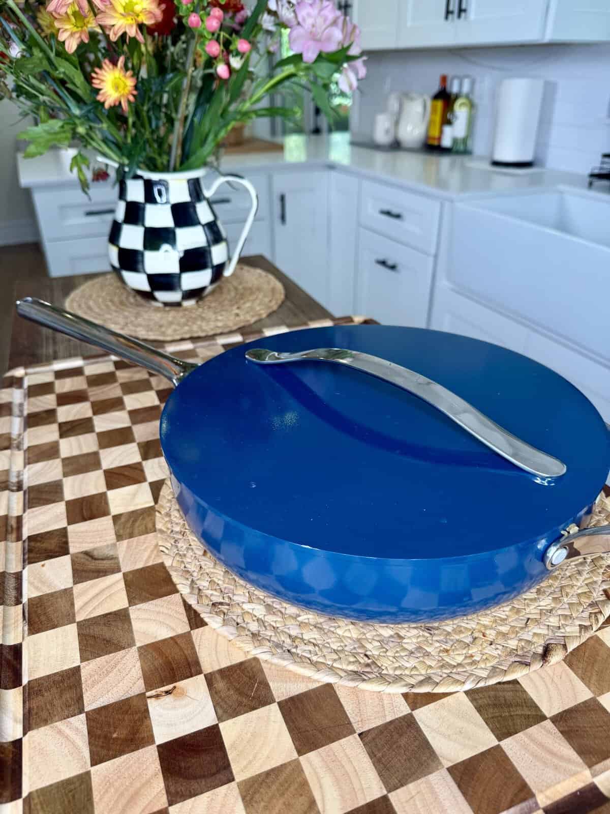 A blue covered pan with a silver handle sits on a woven placemat on a checkered cutting board. Behind it, a black and white checkered vase with colorful flowers is on a kitchen island. White cabinets are in the background.