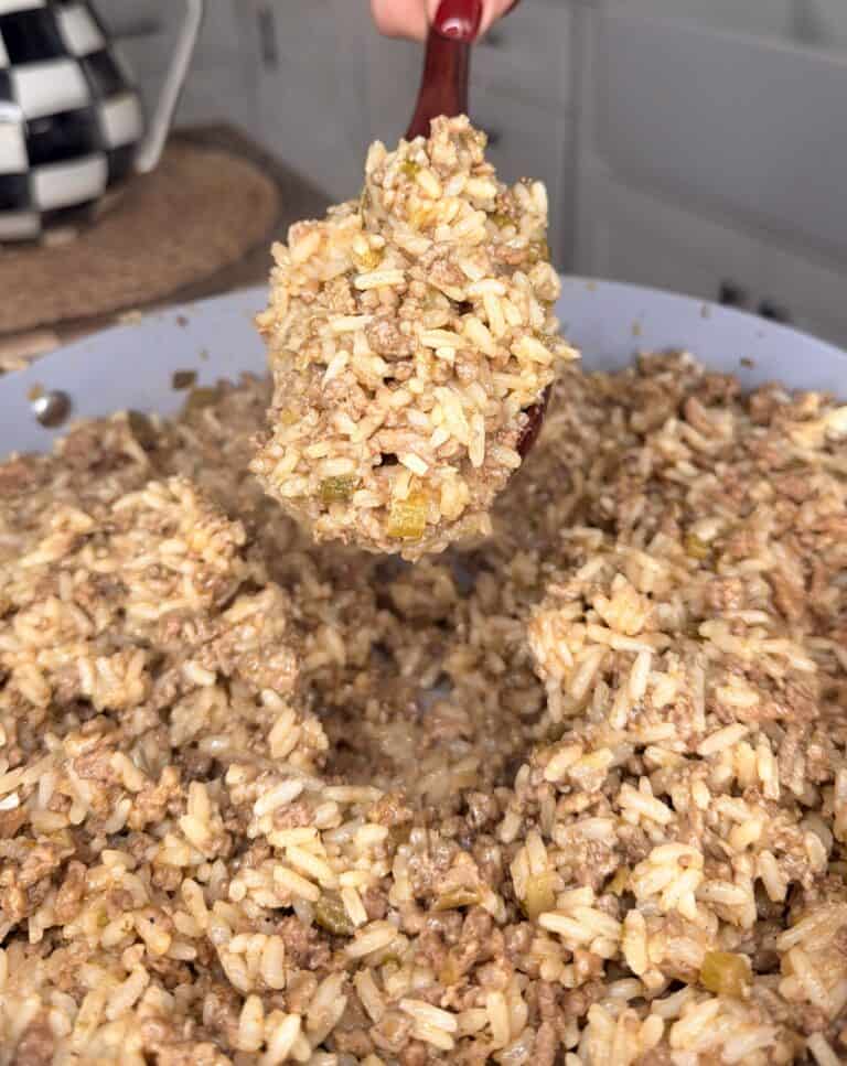 A close-up of a pan filled with cooked rice mixed with ground beef and seasonings. A spoon is lifting a portion from the pan in a kitchen setting.
