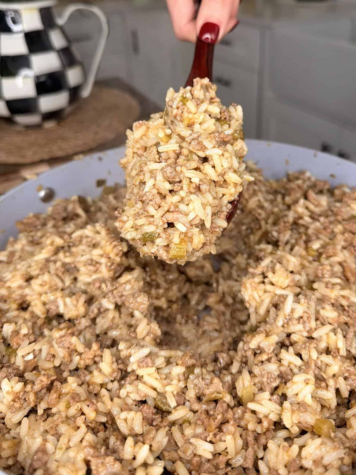 A close-up of a spoonful of seasoned rice mixed with ground beef and vegetables being lifted from a large pan, with a kitchen background visible.