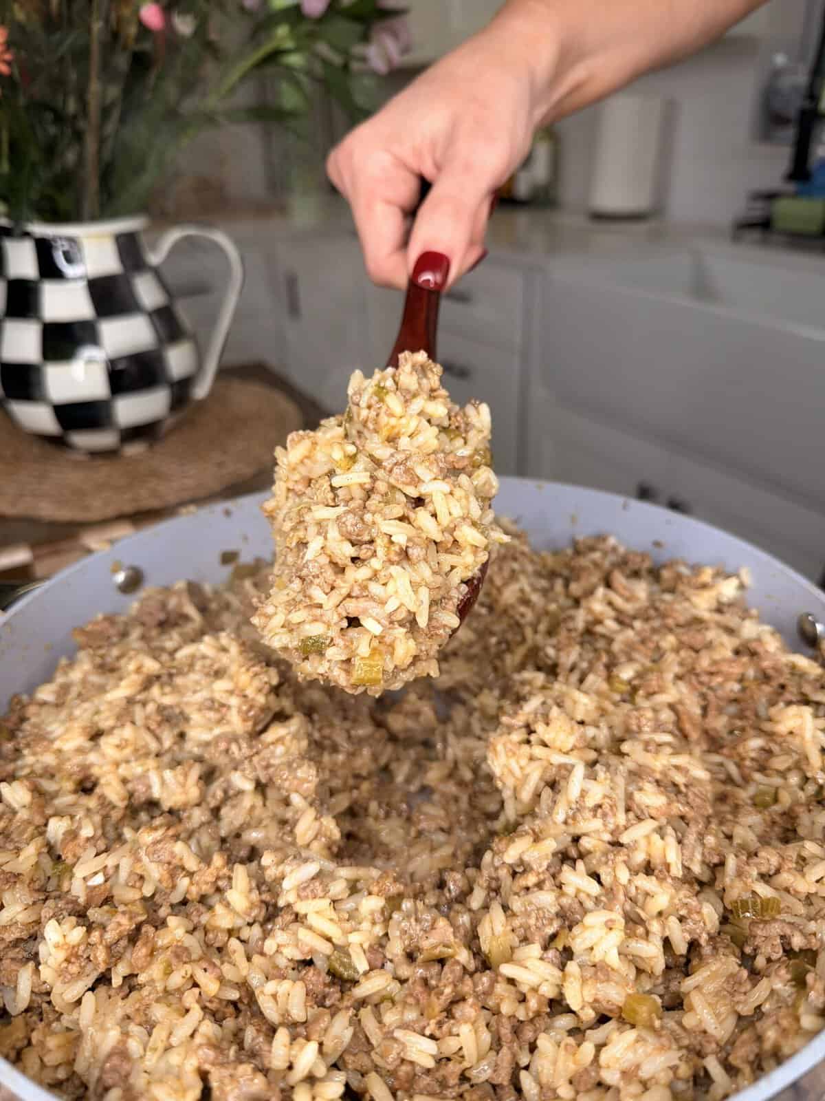 A hand with red nail polish holds a spoonful of rice mixed with ground meat and vegetables over a large pan filled with the same dish in a kitchen setting.