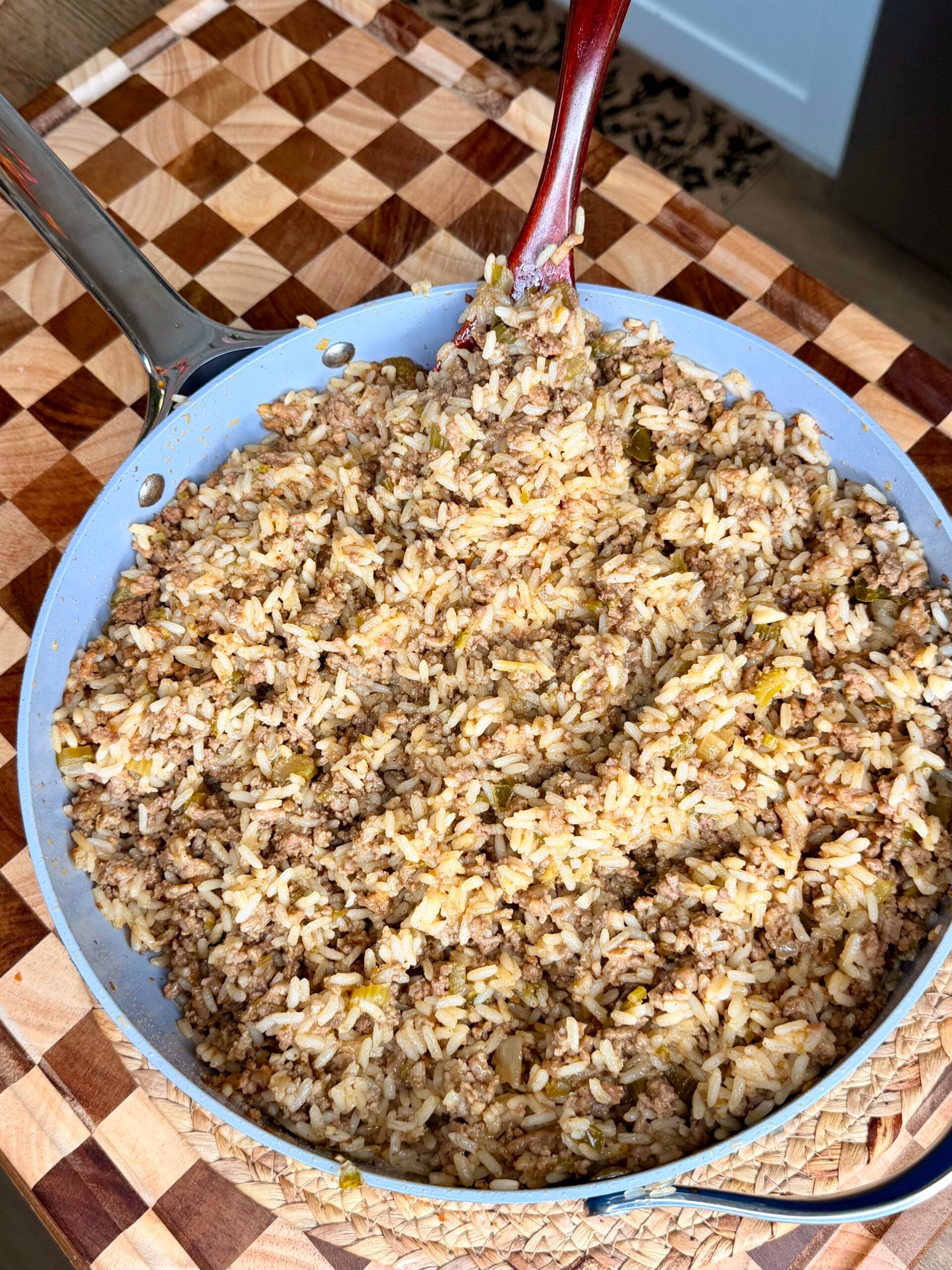 A skillet filled with cooked rice mixed with ground beef and seasonings sits on a checkered wooden cutting board, with a red spoon resting in the pan.