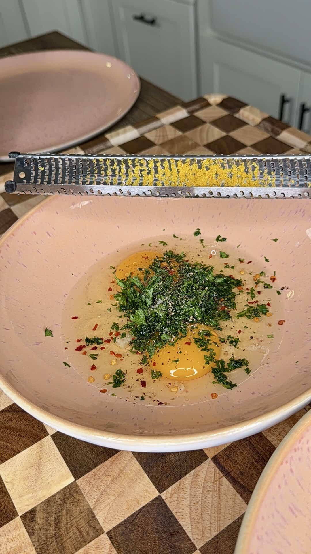 Two cracked eggs in a pink bowl with chopped herbs, pepper, and chili flakes, under a grater topped with lemon zest on a checkered wooden countertop. Pink plates are in the background.
