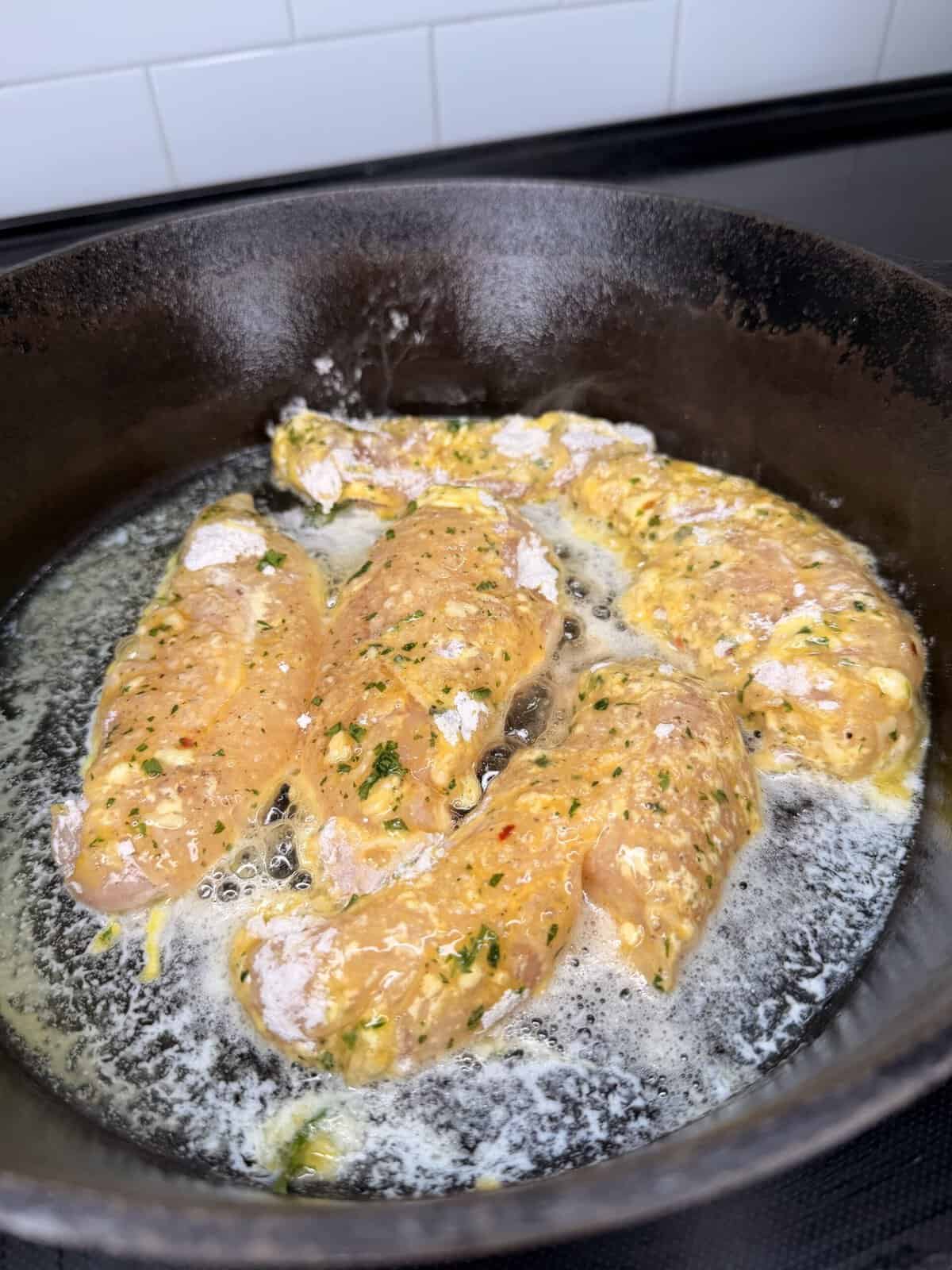 Raw breaded chicken strips frying in a cast iron skillet with bubbling oil, against a white tiled background.