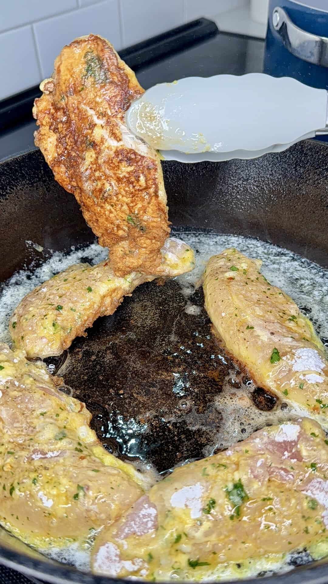 Four pieces of seasoned chicken cook in a cast iron skillet. One piece is being flipped with tongs, showing its golden-brown, crispy side. Butter or oil bubbles in the pan.