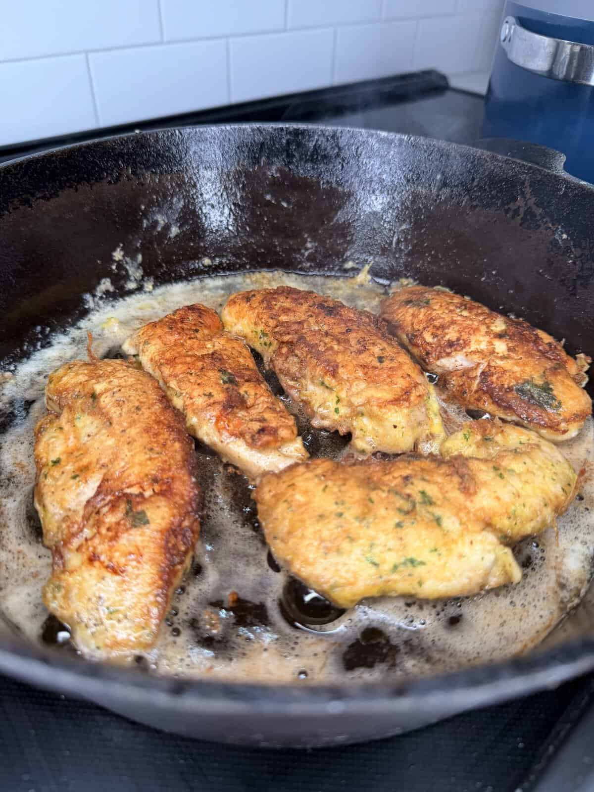 Five golden-brown chicken cutlets are frying in a cast iron skillet with bubbling oil, set on a stovetop in a kitchen with a white tile backsplash.