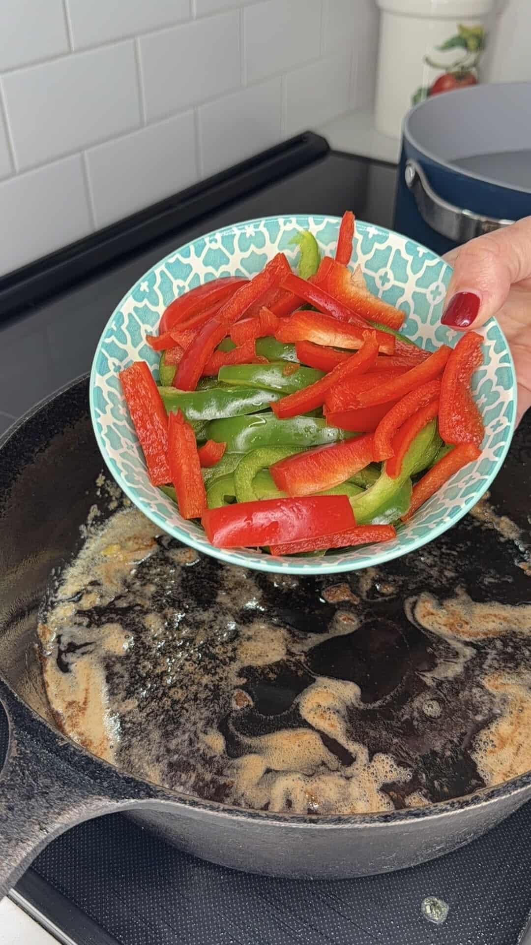 A hand holds a bowl of sliced red and green bell peppers over a cast iron skillet with melted butter on a stovetop, preparing to sautรฉ the vegetables.