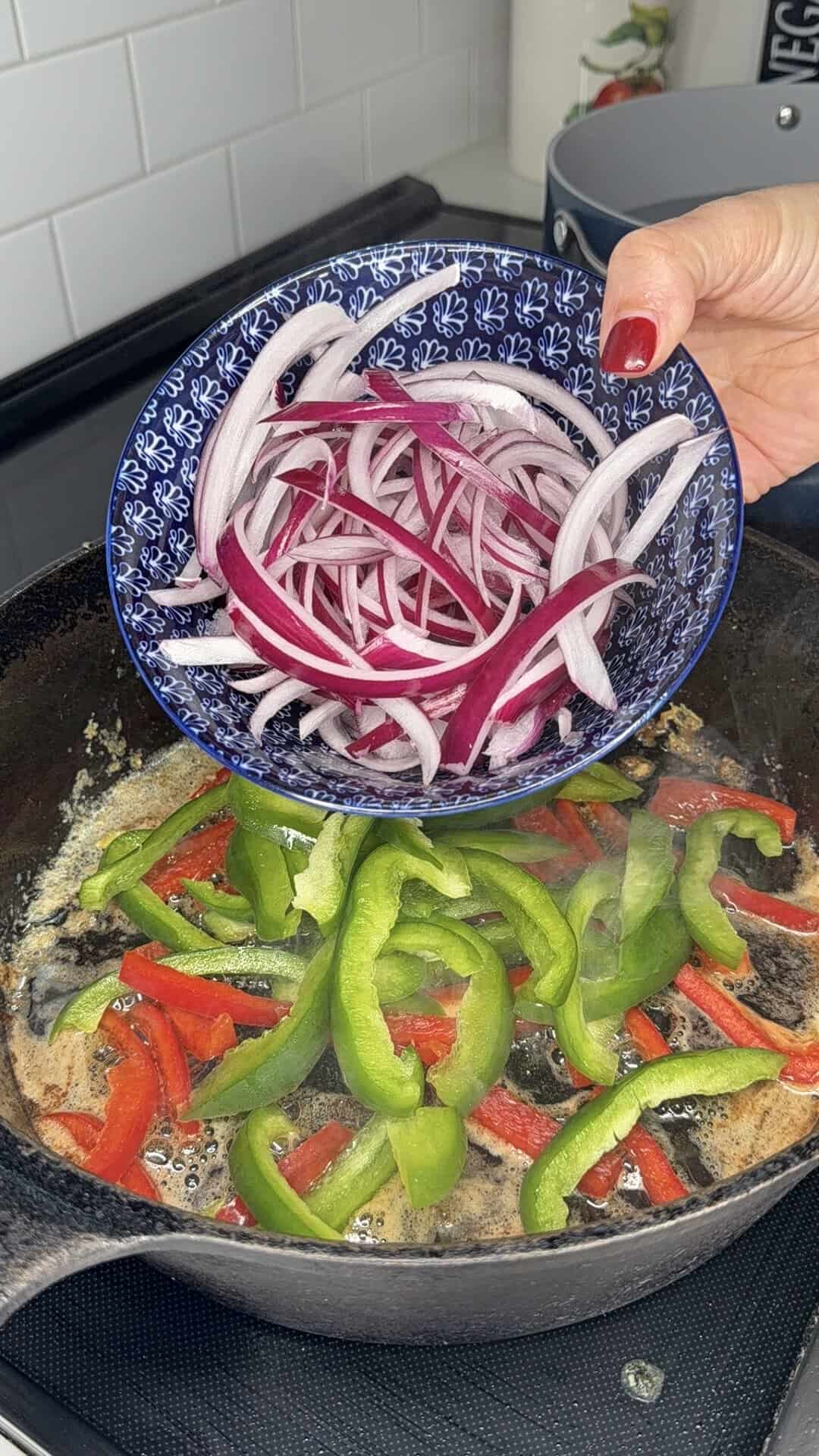 A hand holds a blue bowl of sliced red onions over a skillet with sizzling green and red bell peppers on a stove.