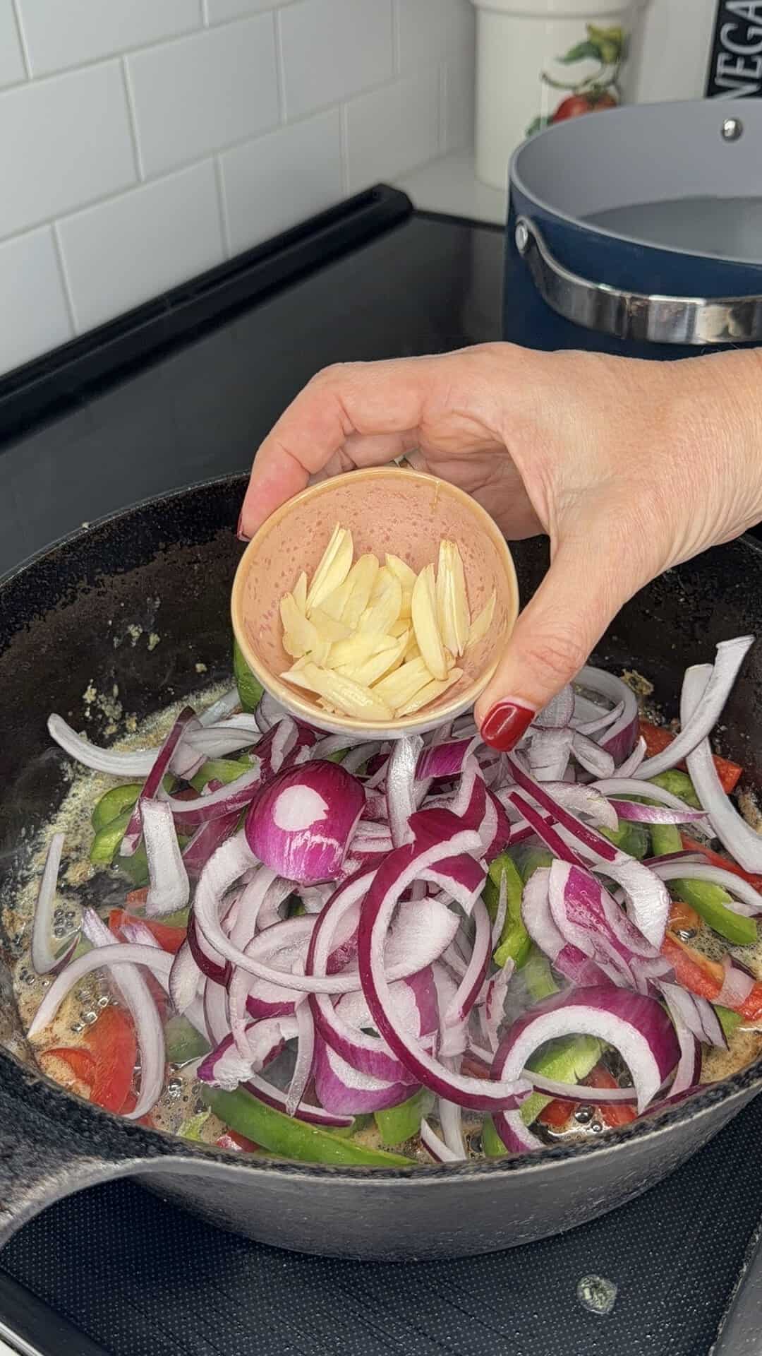 A hand holds a small bowl of sliced garlic above a skillet with sliced red onions, green peppers, and tomatoes cooking on a stovetop.