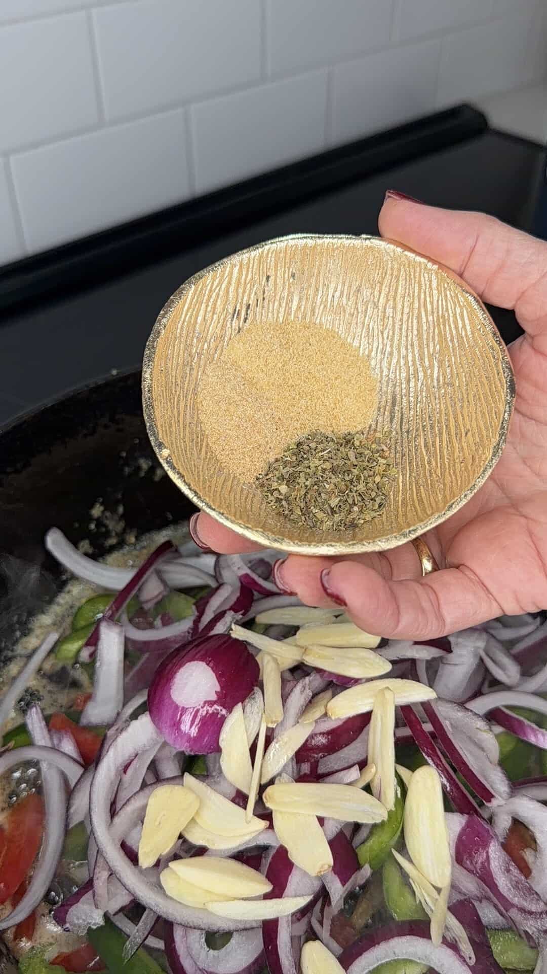 A hand holds a small bowl with seasoning over a pan of sliced red onions, garlic, and vegetables on a stovetop. White tile backsplash is visible in the background.