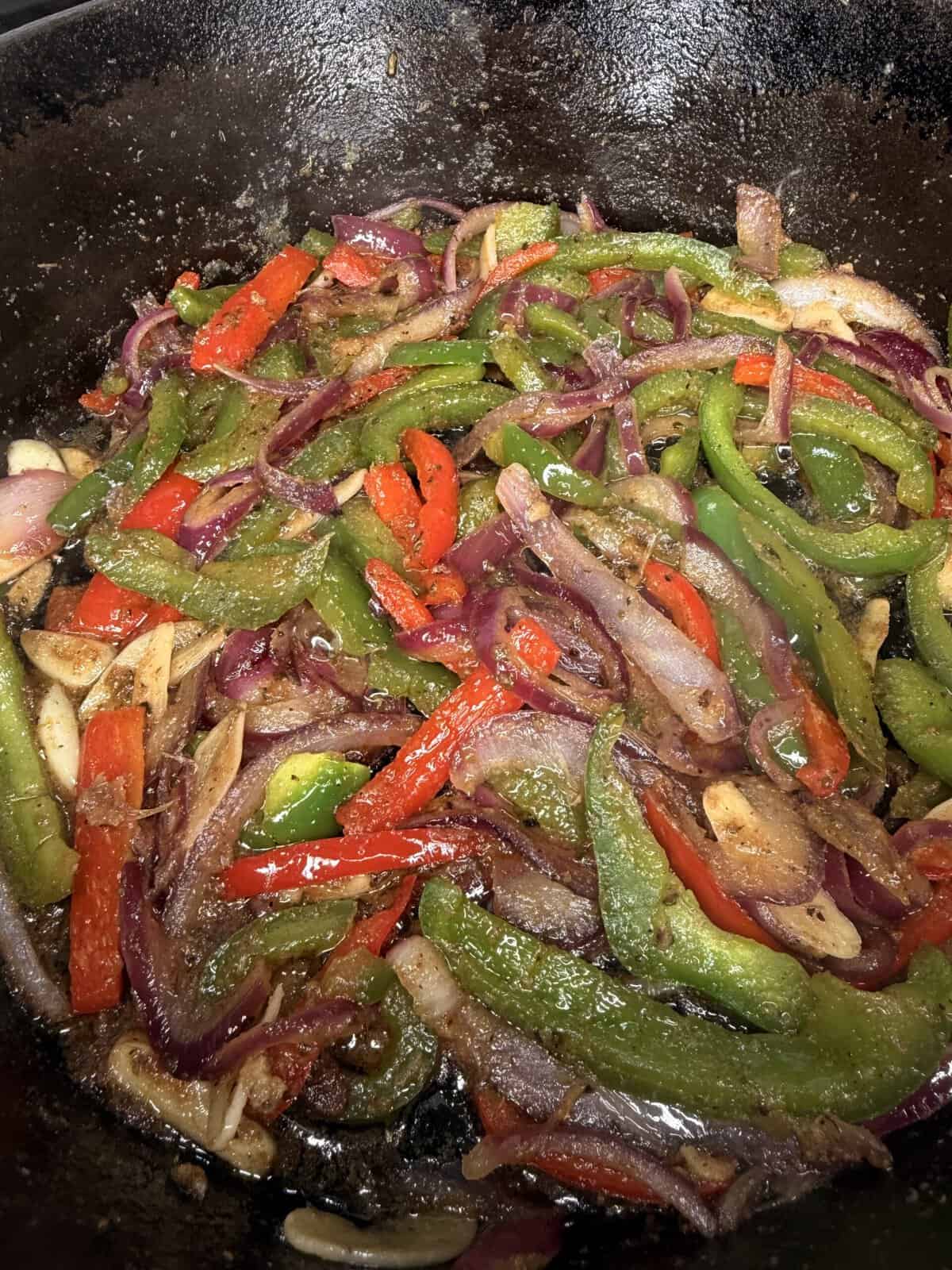 Sliced red and green bell peppers, red onions, and garlic are sautรฉing in a cast iron pan, seasoned with spices.