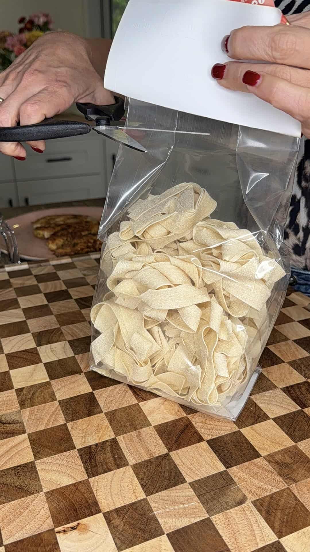 A person with red nail polish uses scissors to cut open a clear plastic bag filled with dry pasta, on a checkered wooden countertop in a kitchen.