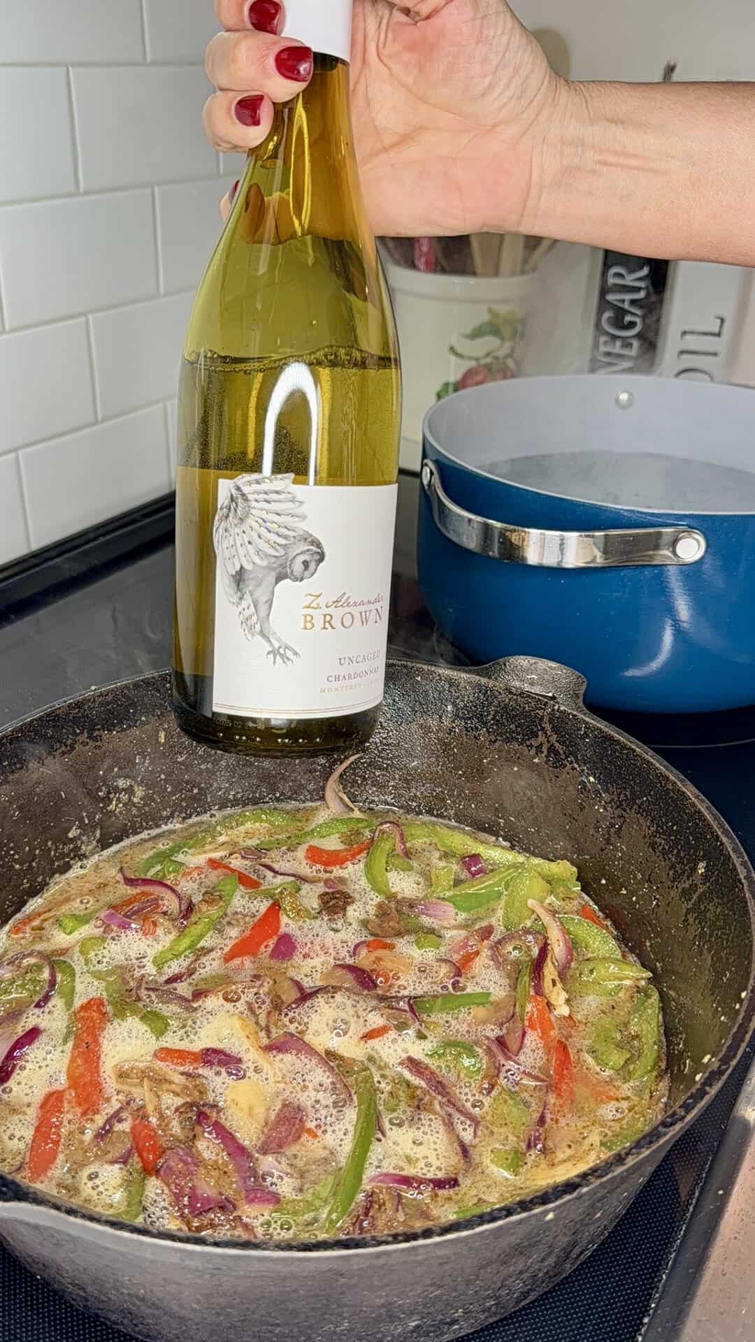 A hand with red nails holds a bottle of white wine over a skillet with simmering sliced peppers and onions; a blue pot on the stove is in the background.