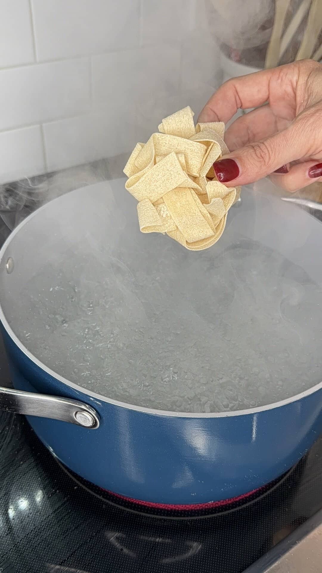 A hand with red nail polish holds a nest of uncooked pasta above a blue pot of boiling water on a stove, with steam rising from the pot.